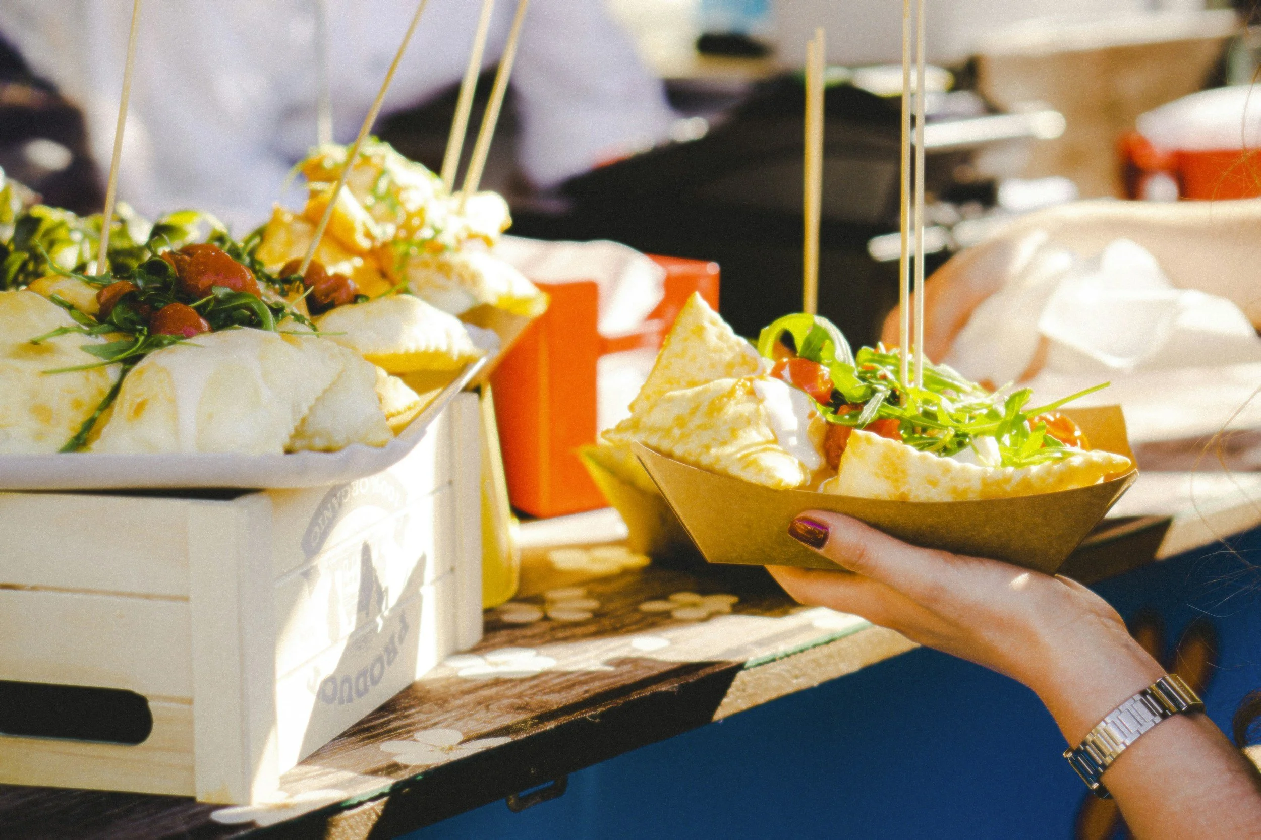 Person holding a bowl of nachos topped with cheese, sour cream, lettuce, tomatoes, and jalapenos at an outdoor food stand, with more nachos on display.