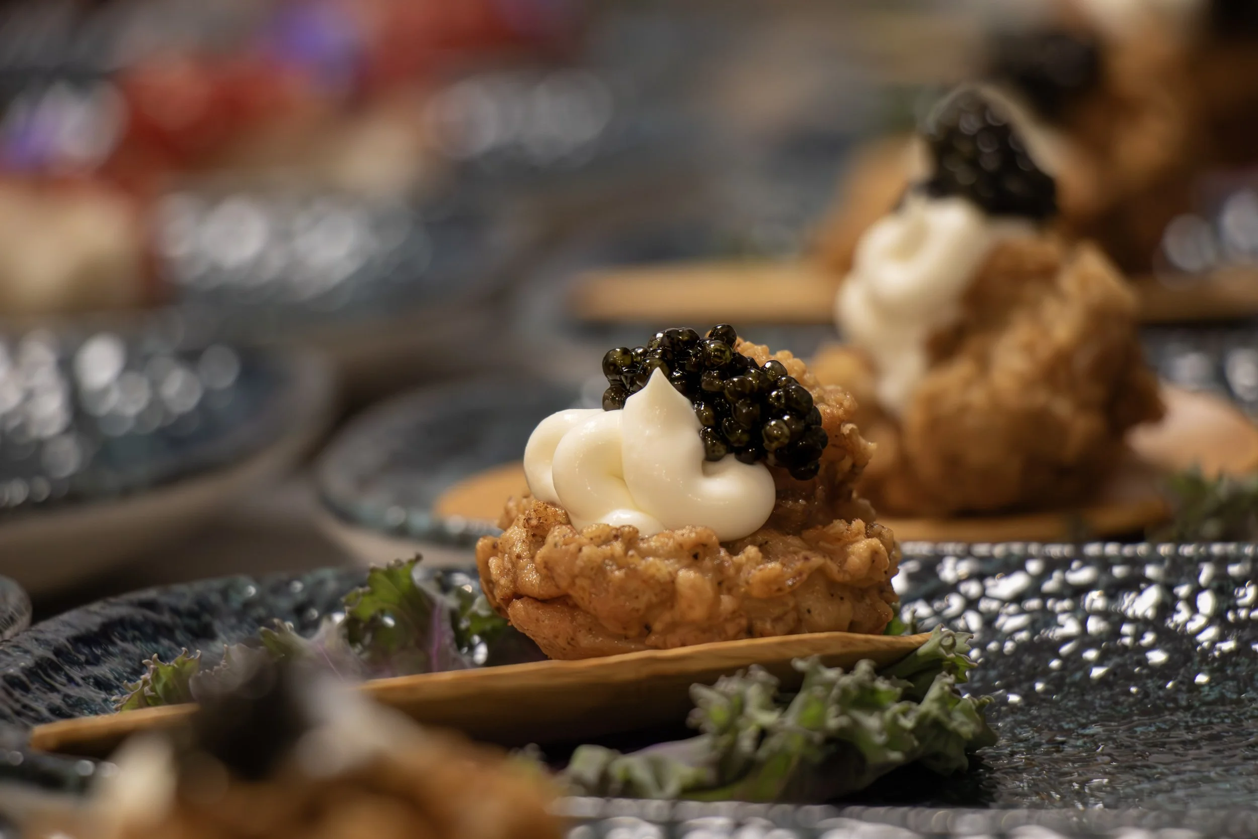 A plated appetizer with a small mound of ground meat, topped with a creamy dollop and black caviar, garnished with green herbs on a decorative plate with a blurred background.