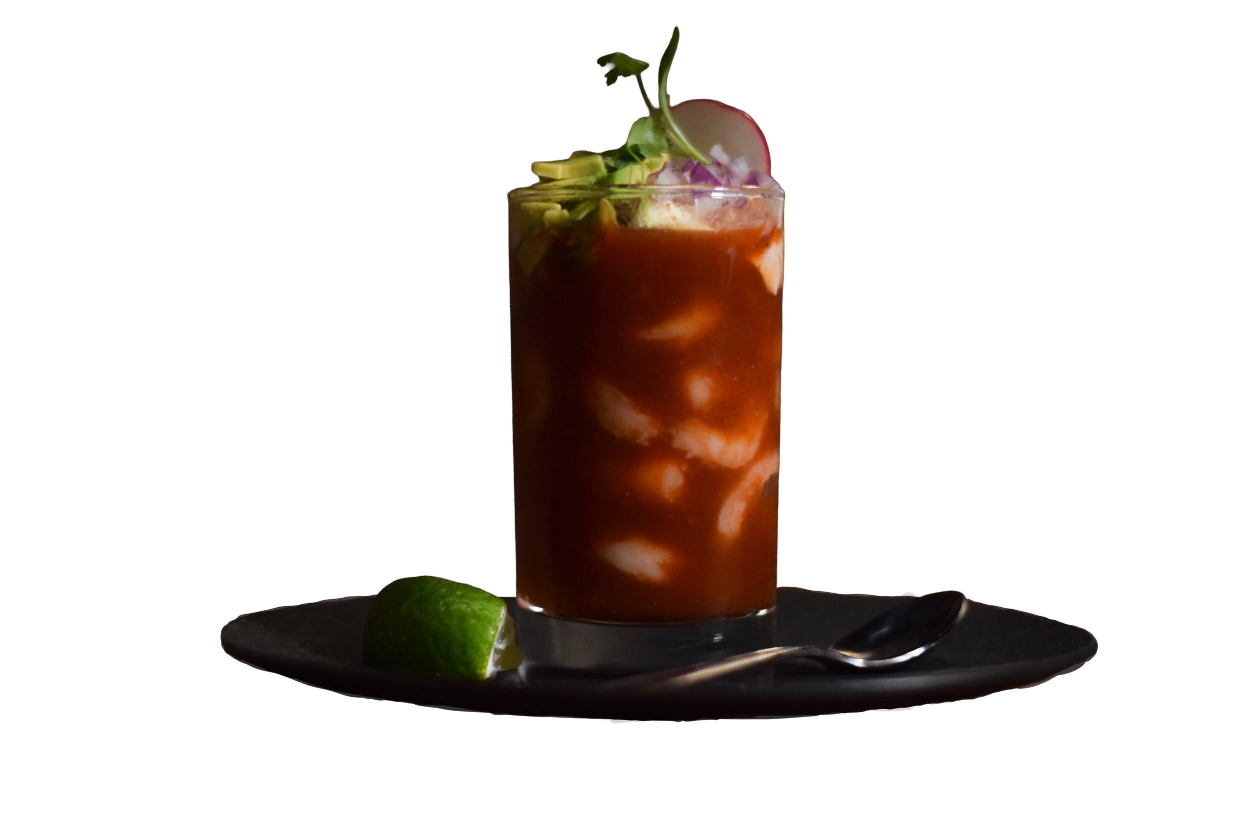 A tall glass of Bloody Mary cocktail garnished with slices of cucumber, radish, and a leafy herb, with lime and spoon next to it on a dark surface.