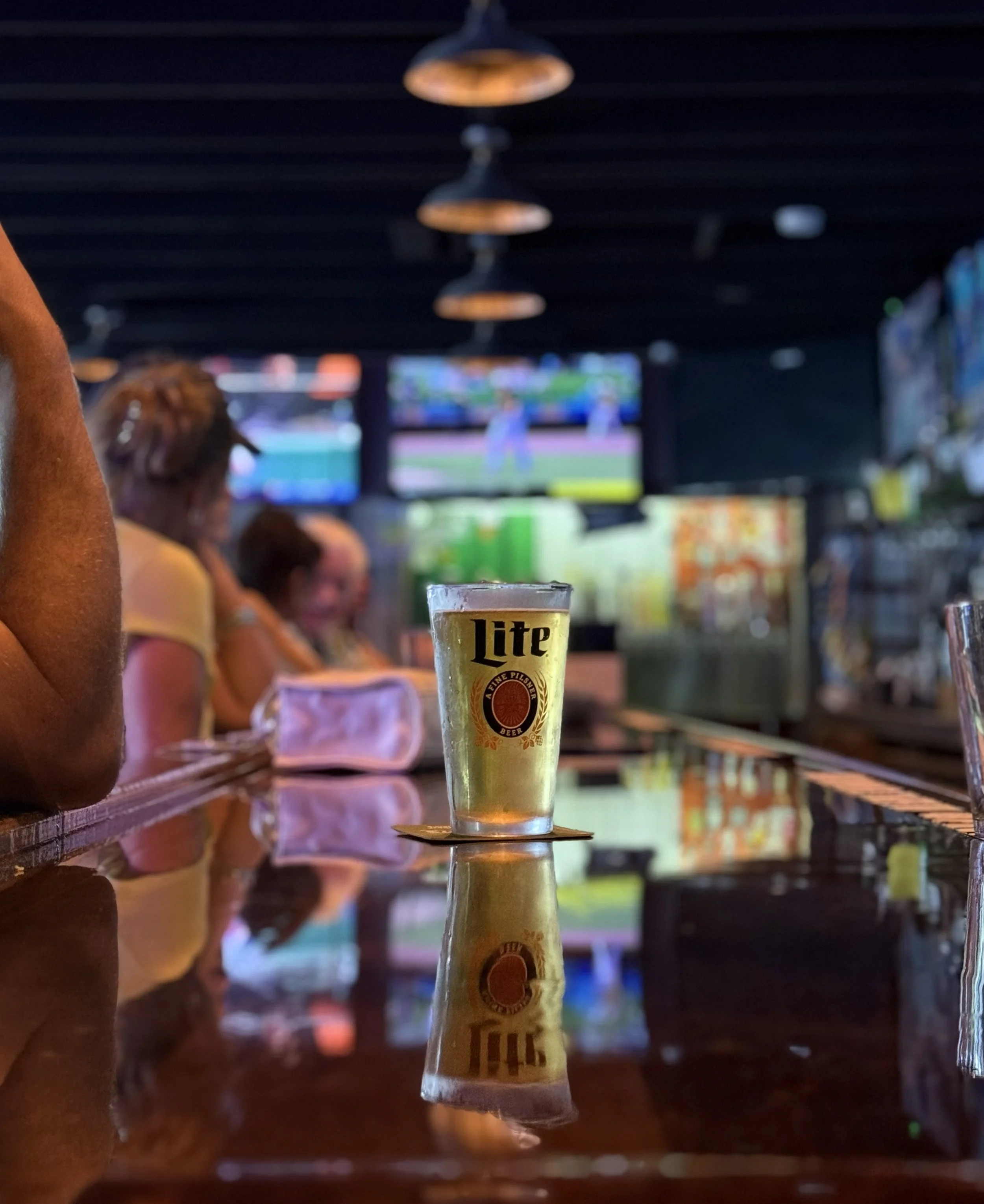 A pint glass of beer on a bar counter with a TV screen in the background showing a baseball game, and people sitting and standing around in a dimly lit bar.