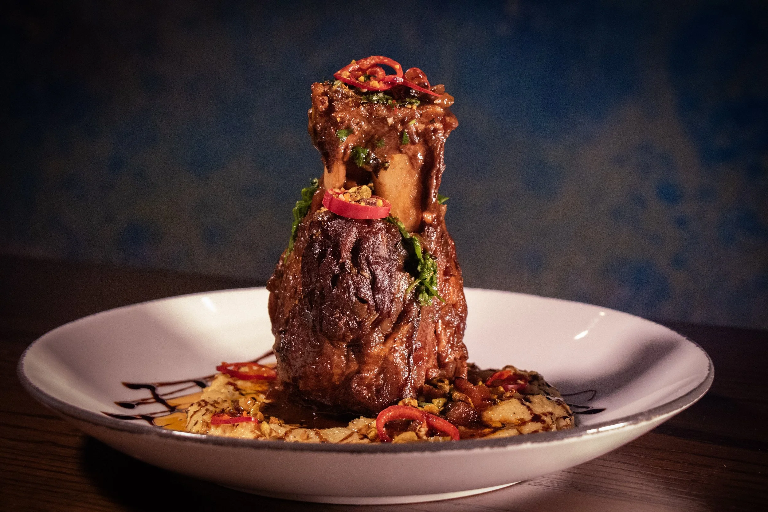 A plated dish featuring a large, glazed meat chop topped with red chili slices and garnished with chopped herbs, accompanied by a colorful vegetable and sauce garnish.