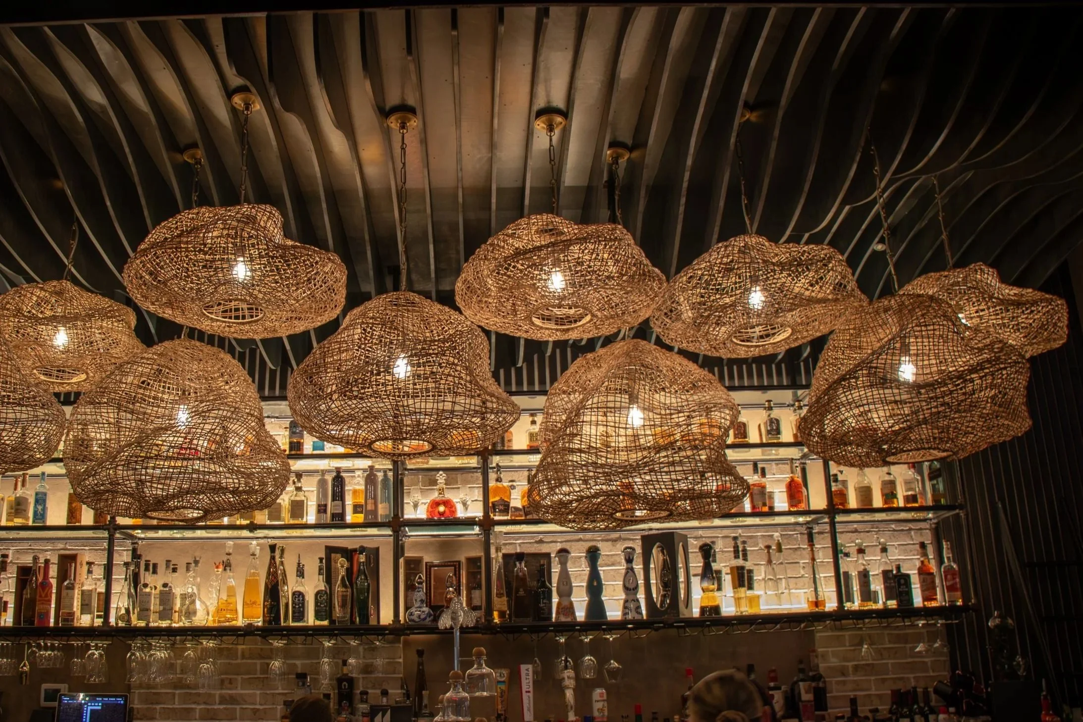 Multiple woven pendant lights hanging above a bar filled with liquor bottles. Audience members are visible at the bottom of the image.