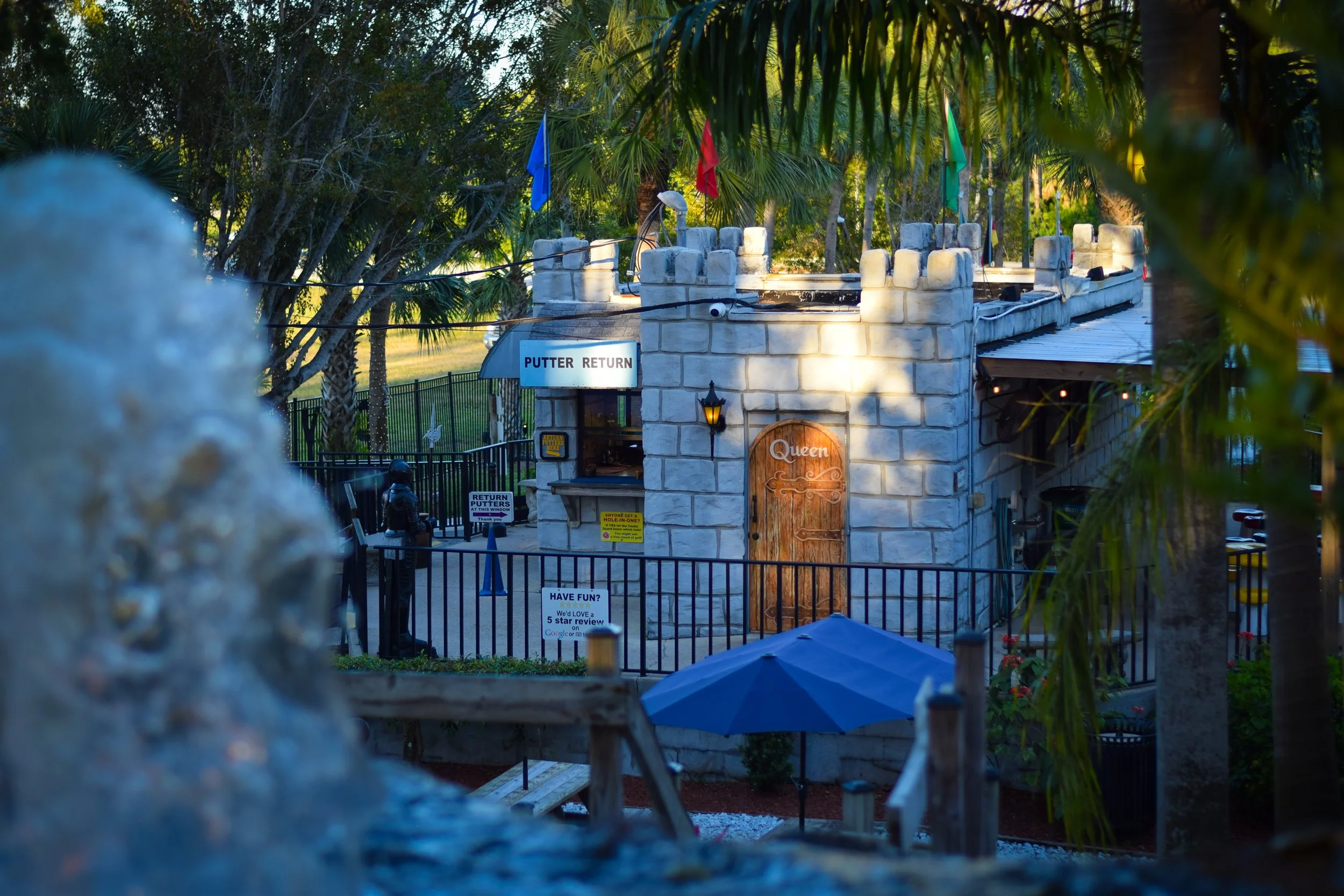 A mini golf course with castle-themed structures, flags, and a sign that reads 'PUTTER RETURN.' There are trees, fences, and umbrellas visible in the background, with a person near the course and various signs for the game.