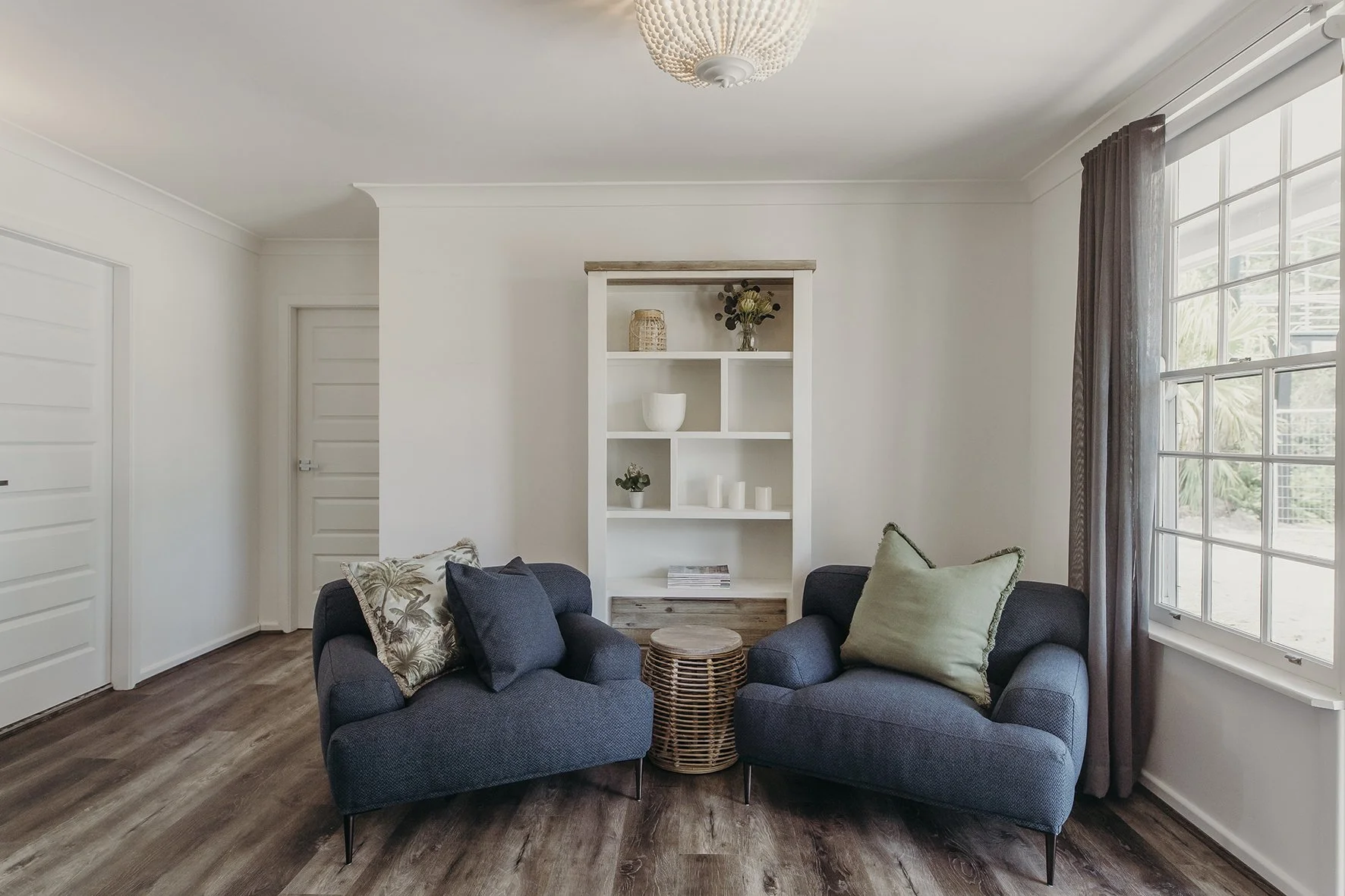 Living room with two dark gray armchairs, a round wicker side table, white walls, a large window with brown curtains, and a white bookshelf with decorative items.