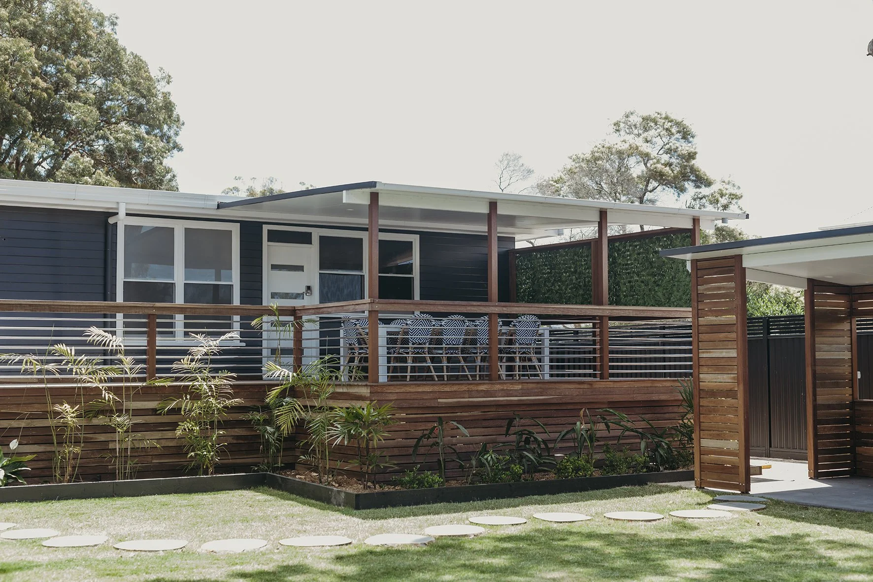 Modern house with a wooden deck, outdoor dining table with chairs, and a small garden with plants and stepping stones.