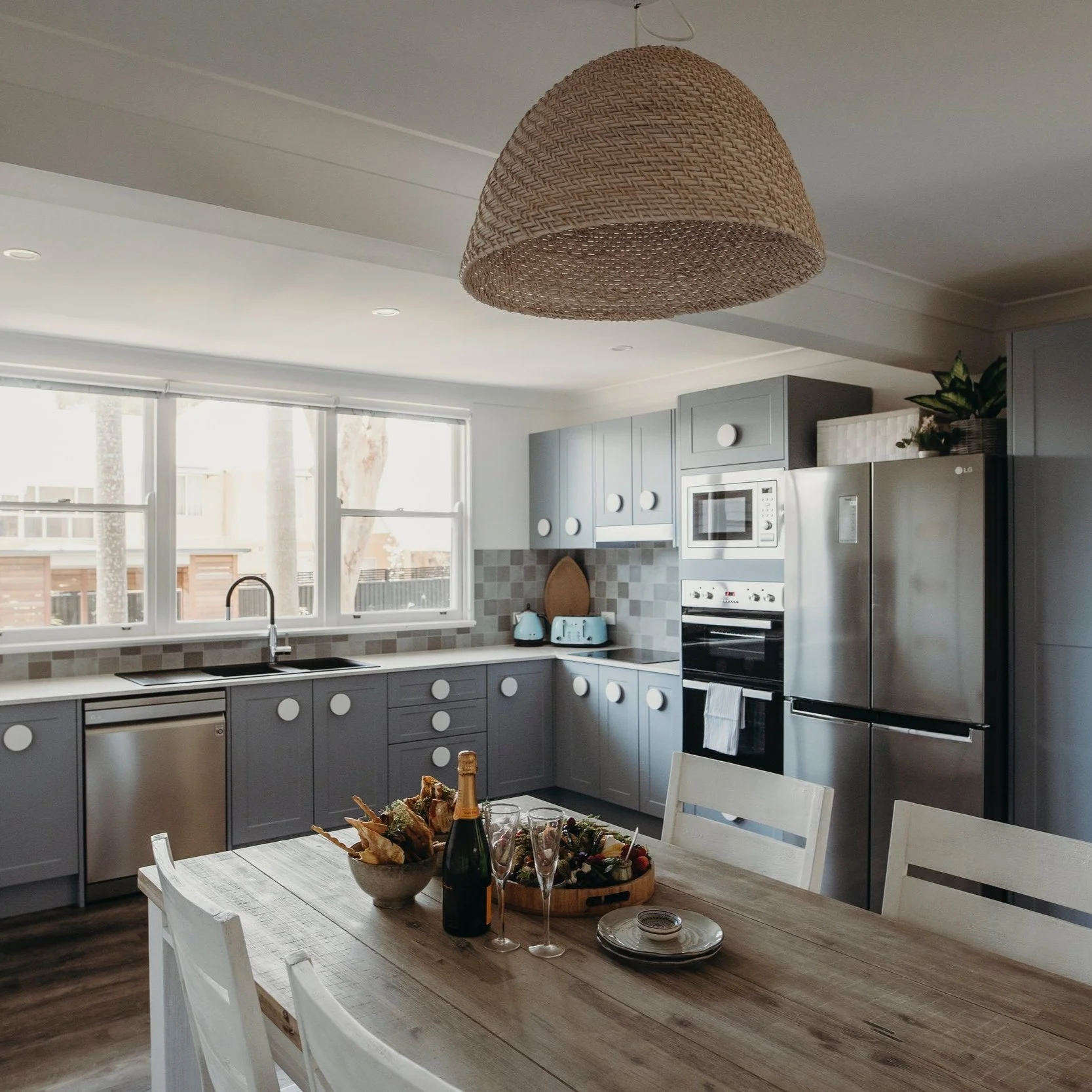 Modern kitchen with gray cabinets, stainless steel refrigerator, oven, microwave, and light gray tiled backsplash. A wooden dining table with plates, a bottle of champagne, and food, near white chairs. Large windows with sunlight, a woven pendant lig