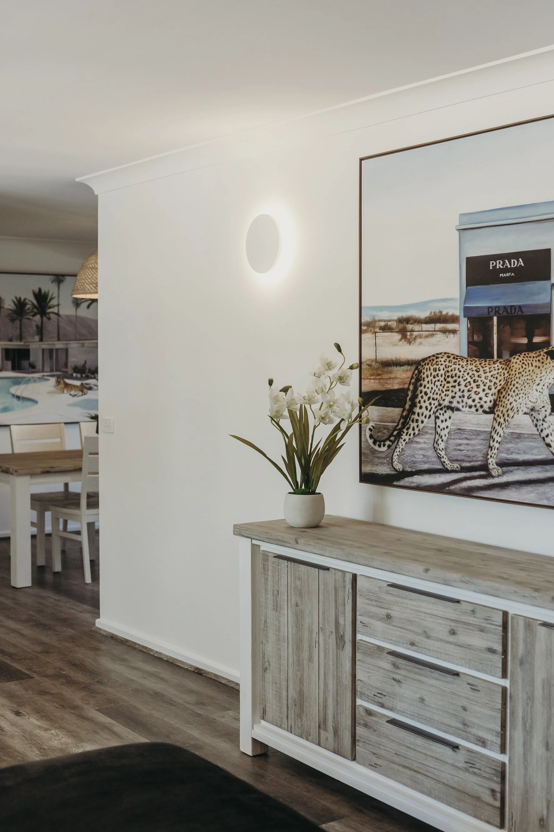 Living room with a white wall, a wooden sideboard, a white vase with white flowers, a wall lamp, and a painting of a leopard walking past Prada storefront.