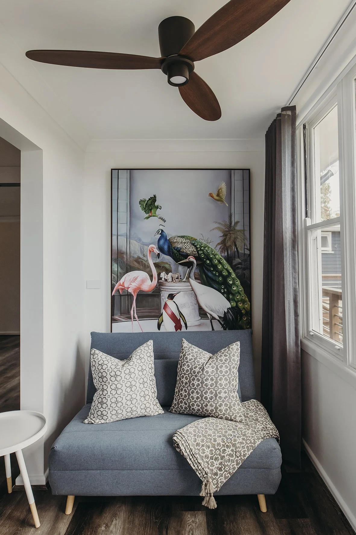 A cozy living room corner with a gray loveseat, two patterned pillows, a patterned blanket, and a large framed artwork of colorful exotic birds behind it. A window with sheer curtains is on the right, and a ceiling fan with wooden blades is on the ce