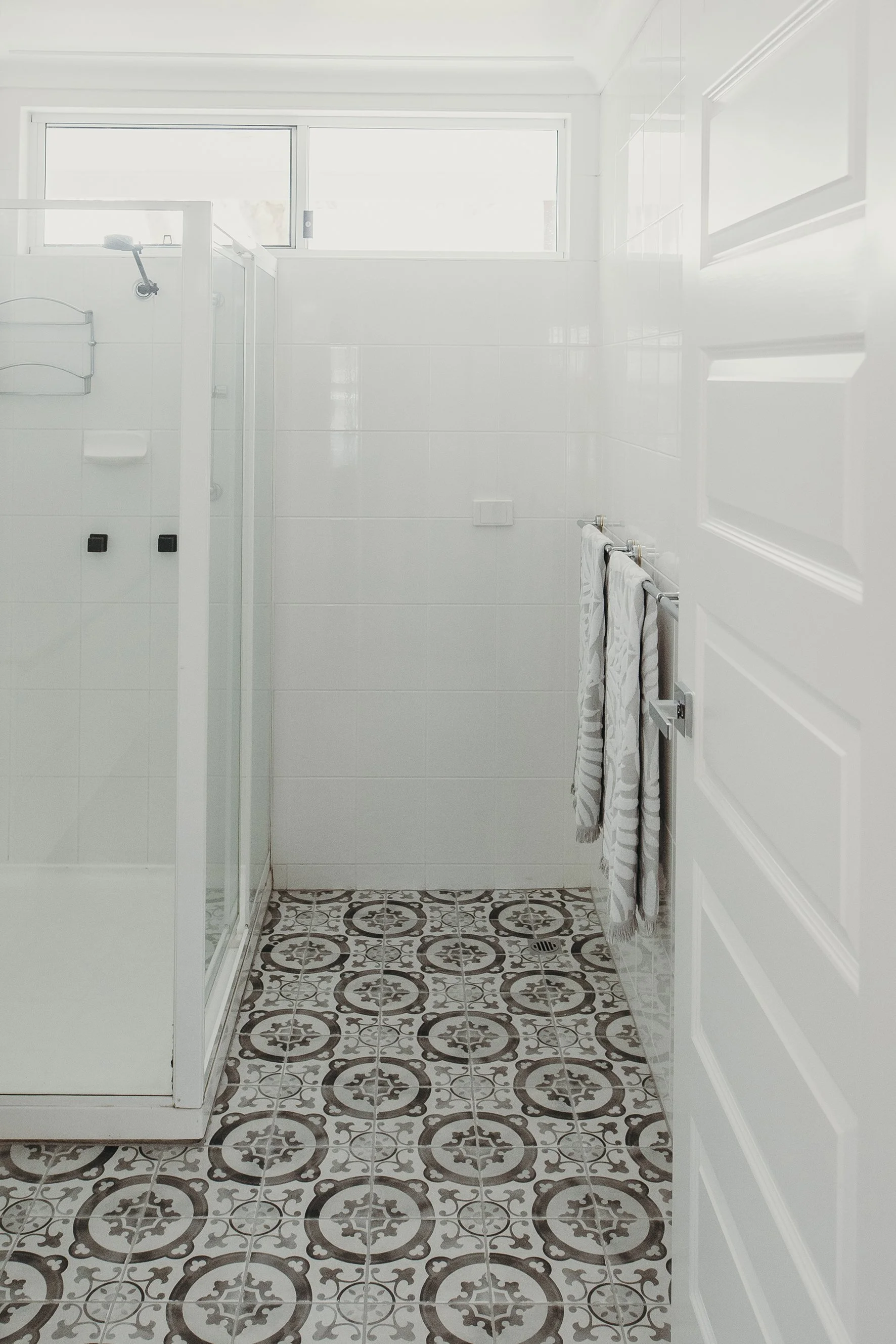 A bright, white bathroom with a glass shower enclosure, white tiles, and patterned floor tiles. A towel rack with two white towels hangs on the right side of the bathroom.