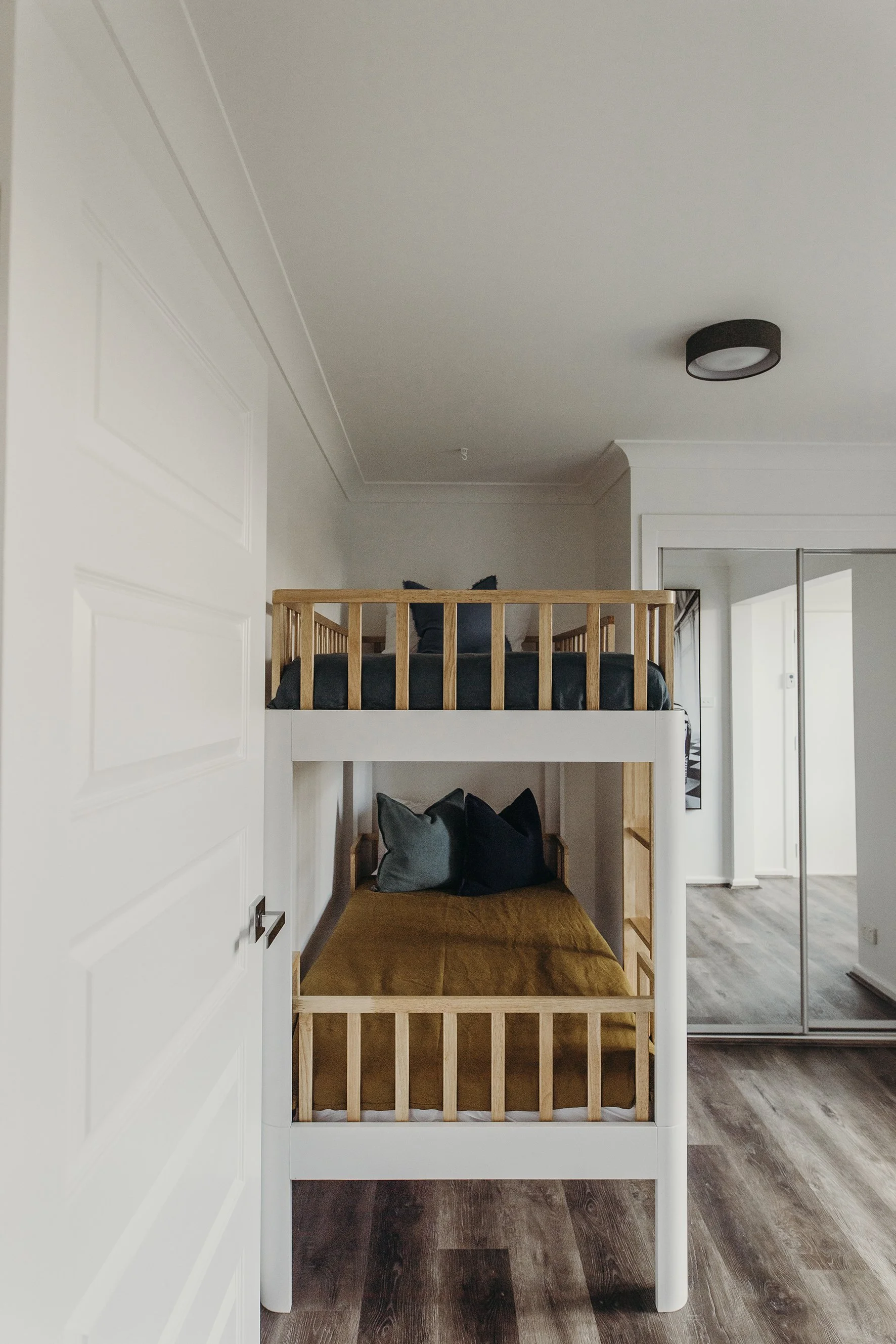 A room with a bunk bed that has a white frame and wooden guardrails on the top bunk. The beds are decorated with pillows, with the bottom bunk having a mustard-colored blanket and two pillows, one grey and one black. There is a closet with mirrored s
