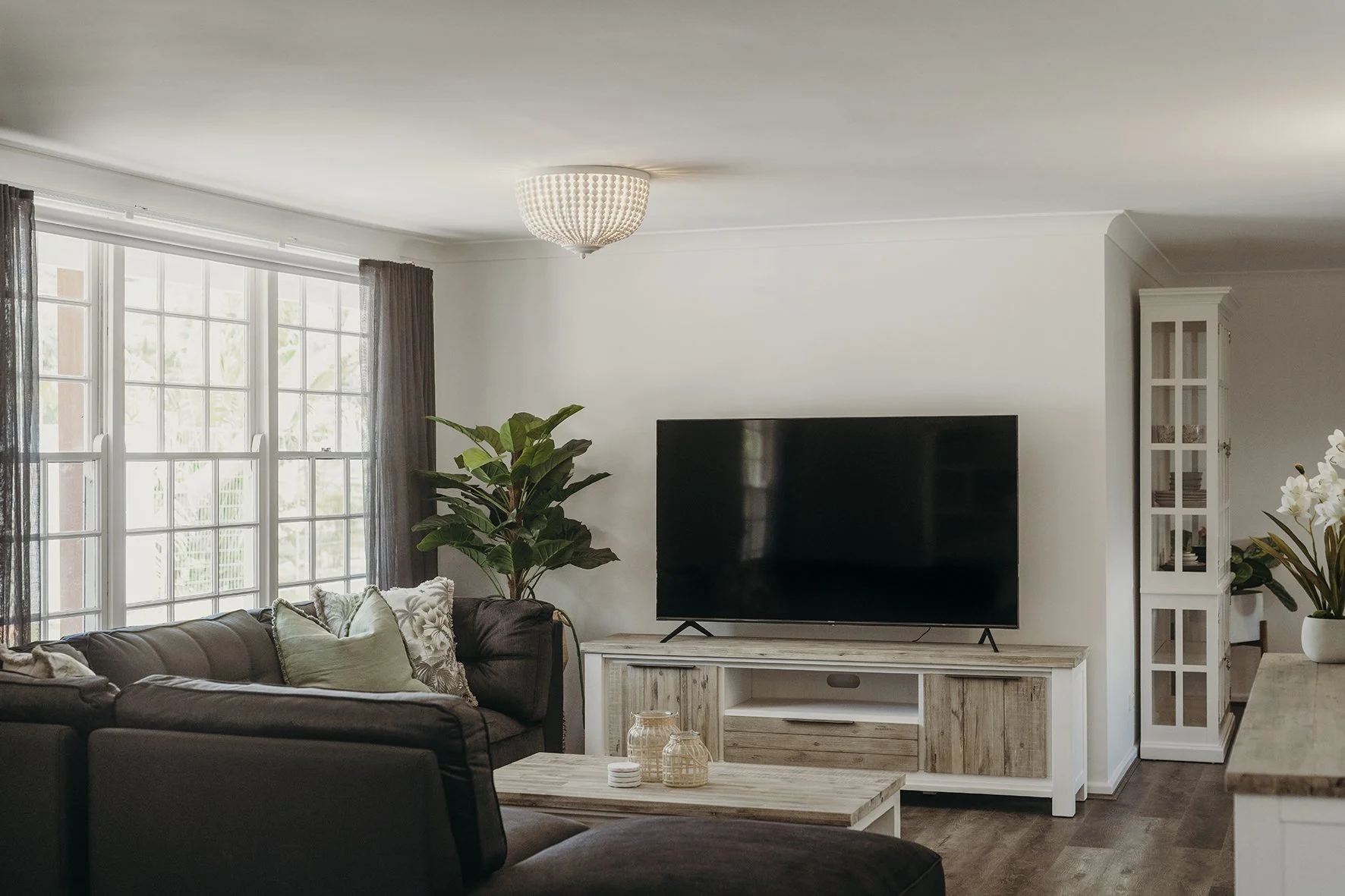 Living room with a large flat-screen TV on a white wooden console, a brown sofa with pillows, a coffee table with decorative jars, a tall green plant, large windows with dark curtains, a white ceiling light, and a white cabinet with glass doors in th
