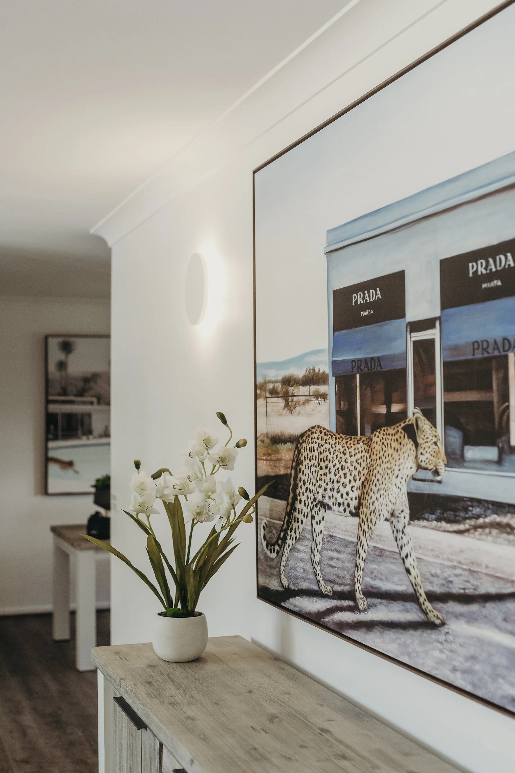 Interior of a home with a wood console table, a white flower vase with orchids in front of a large framed painting of a jaguar in front of a Prada storefront, and additional art in the background.