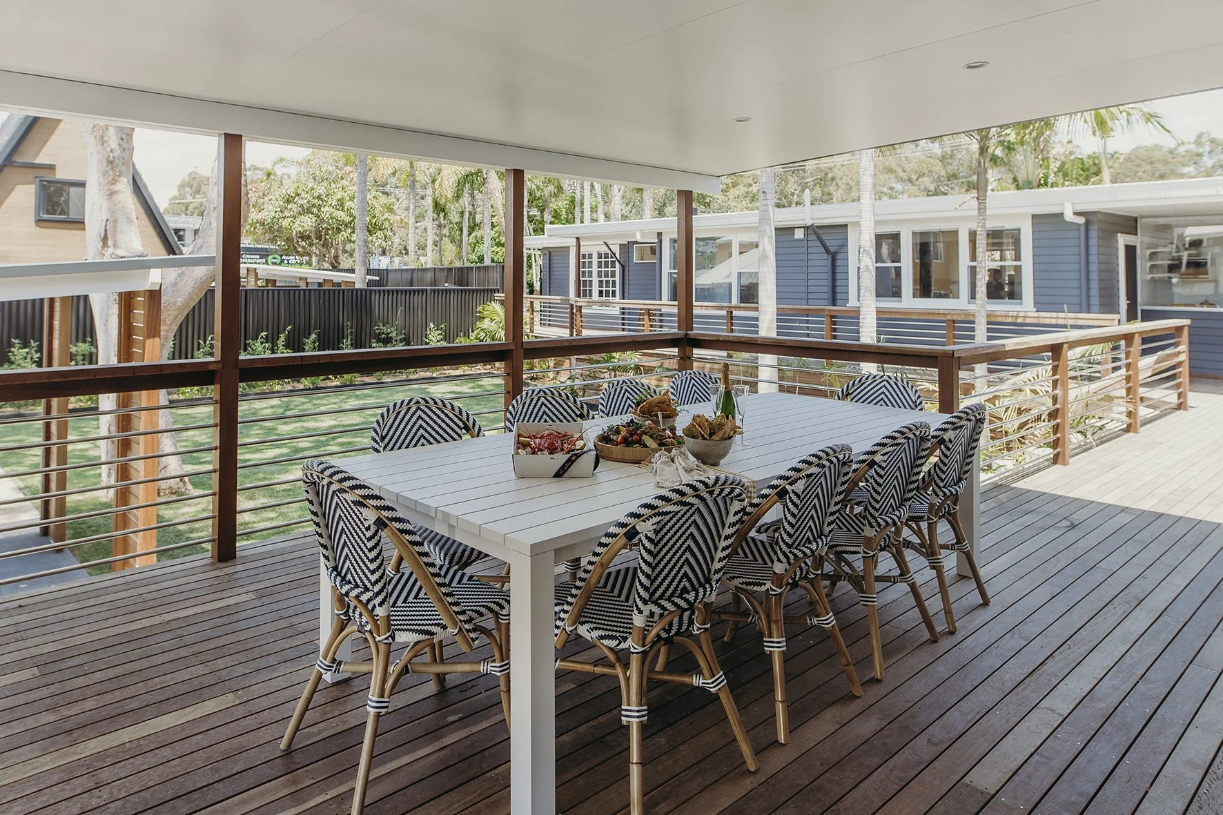 Outdoor patio with a white dining table and ten patterned chairs, set with food and drinks, overlooking a backyard with trees and neighboring houses.