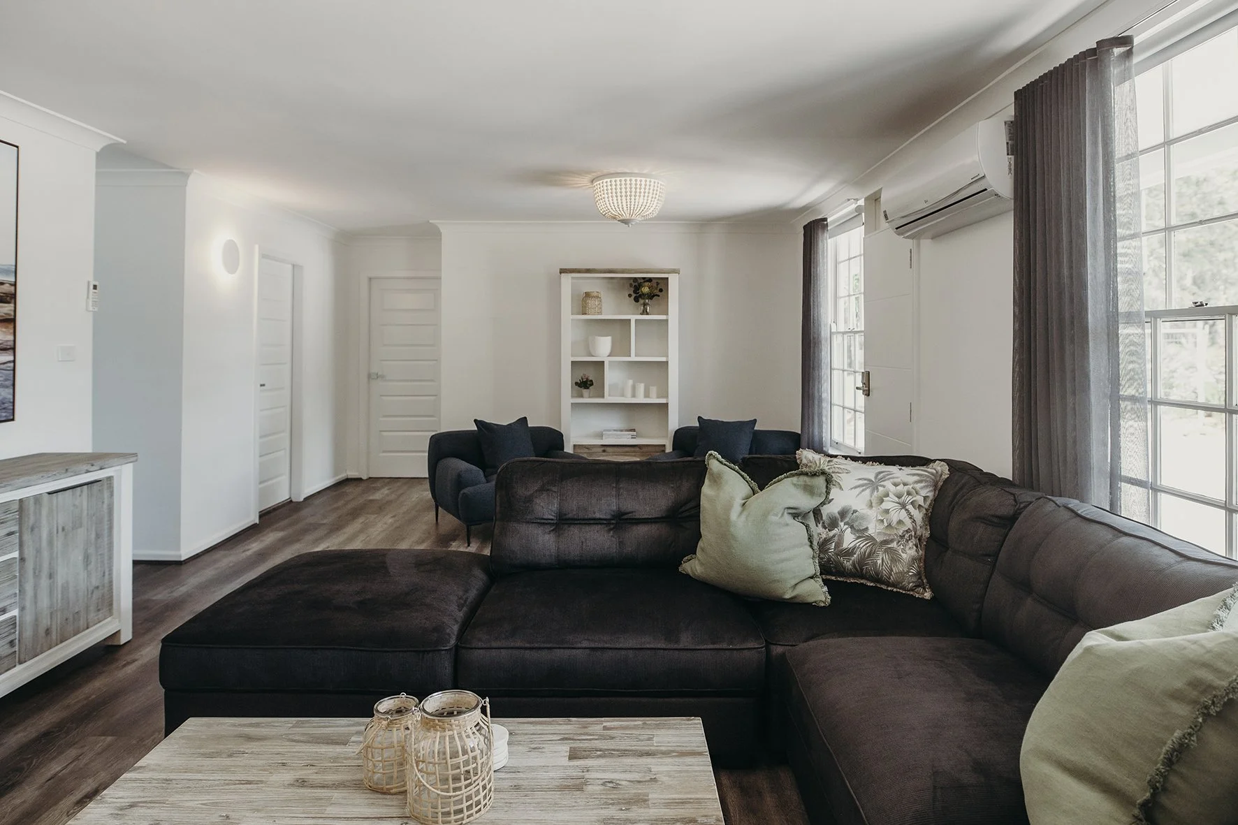Living room with brown sectional sofa, throw pillows, wooden coffee table with decorative lanterns, white shelves, dark curtains, windows, and air conditioning unit.