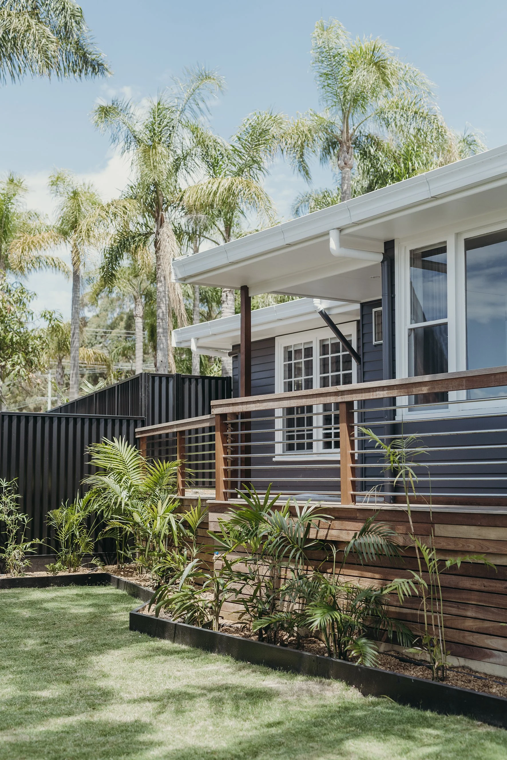 Backyard with a wooden deck, grass lawn, tropical plants, trees, and a house with dark blue exterior, large windows, and a white roof.