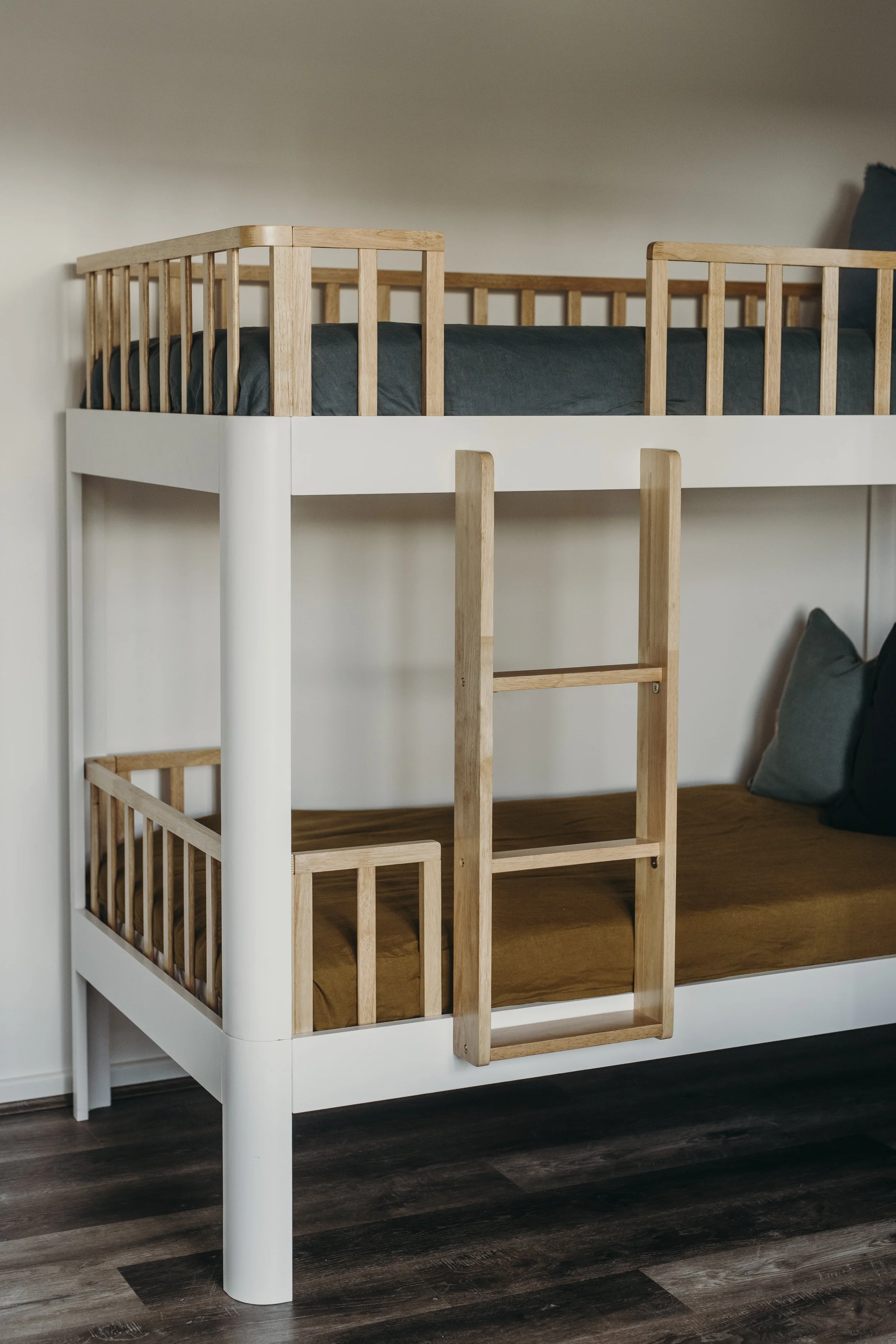A white bunk bed with wooden railings and ladder, with two mattresses and pillows, in a room with dark wood flooring.