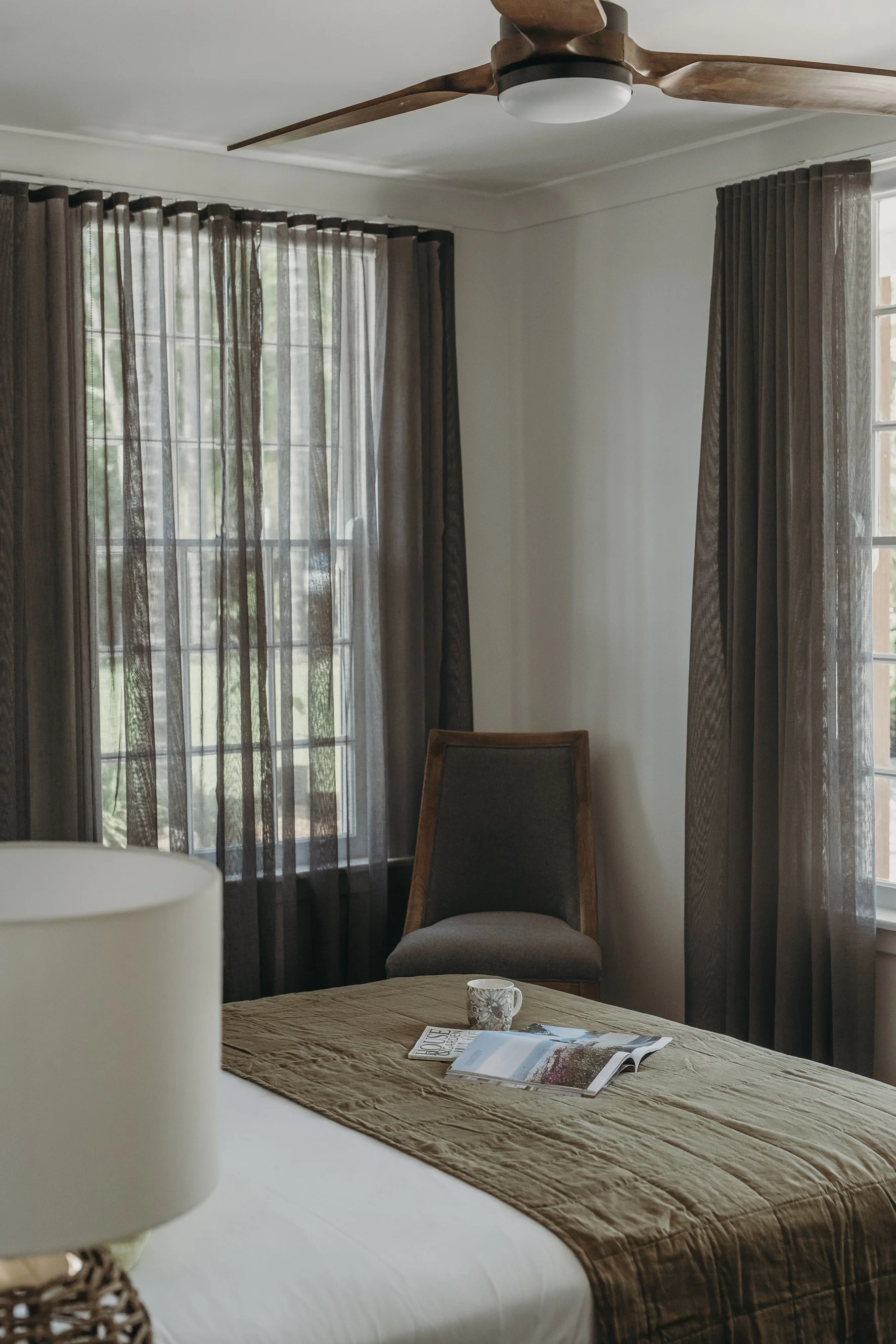 A bedroom with brown curtains on the windows, a table with a magazine and mug, a chair, and a ceiling fan.