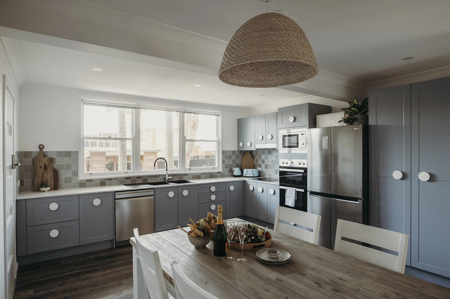 Modern kitchen with gray cabinets, stainless steel appliances, a wooden dining table set with a champagne bottle, glasses, and dishes, and a large woven pendant light overhead.