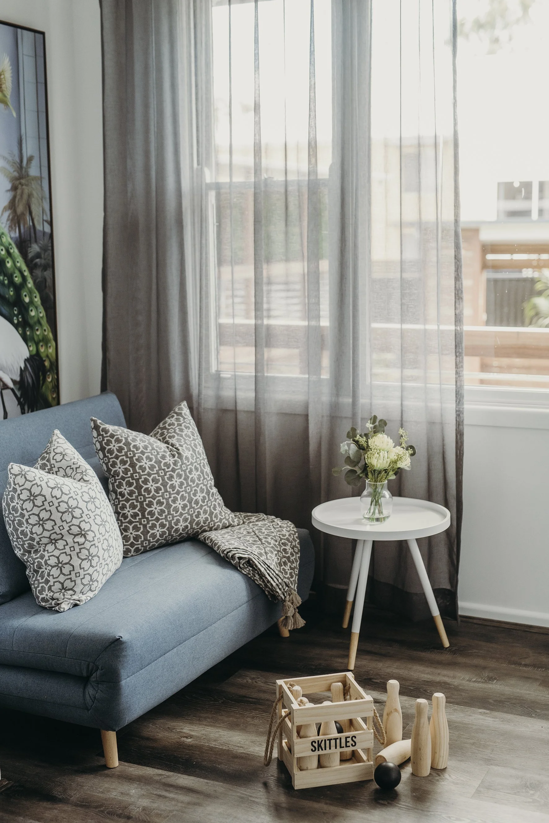 Living room corner with a blue couch, four patterned pillows, a blanket, a small white side table with a vase of white flowers, and a wooden bowling set labeled 'SKITTLES' on the floor, near a window with sheer curtains.