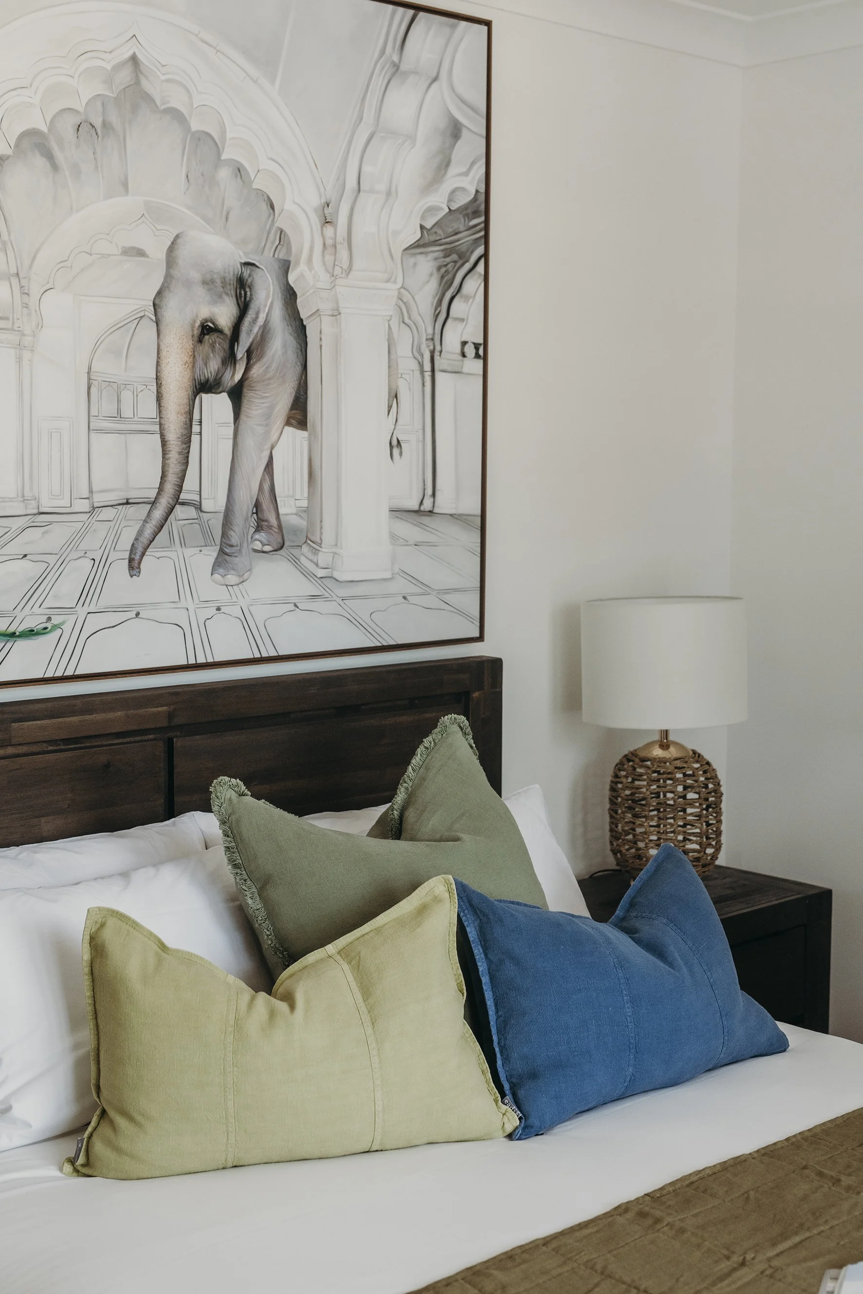 Bedroom with a large framed art of an elephant, multiple pillows on the bed in shades of yellow, green, and blue, and a wicker-based lamp on a dark wooden bedside table.
