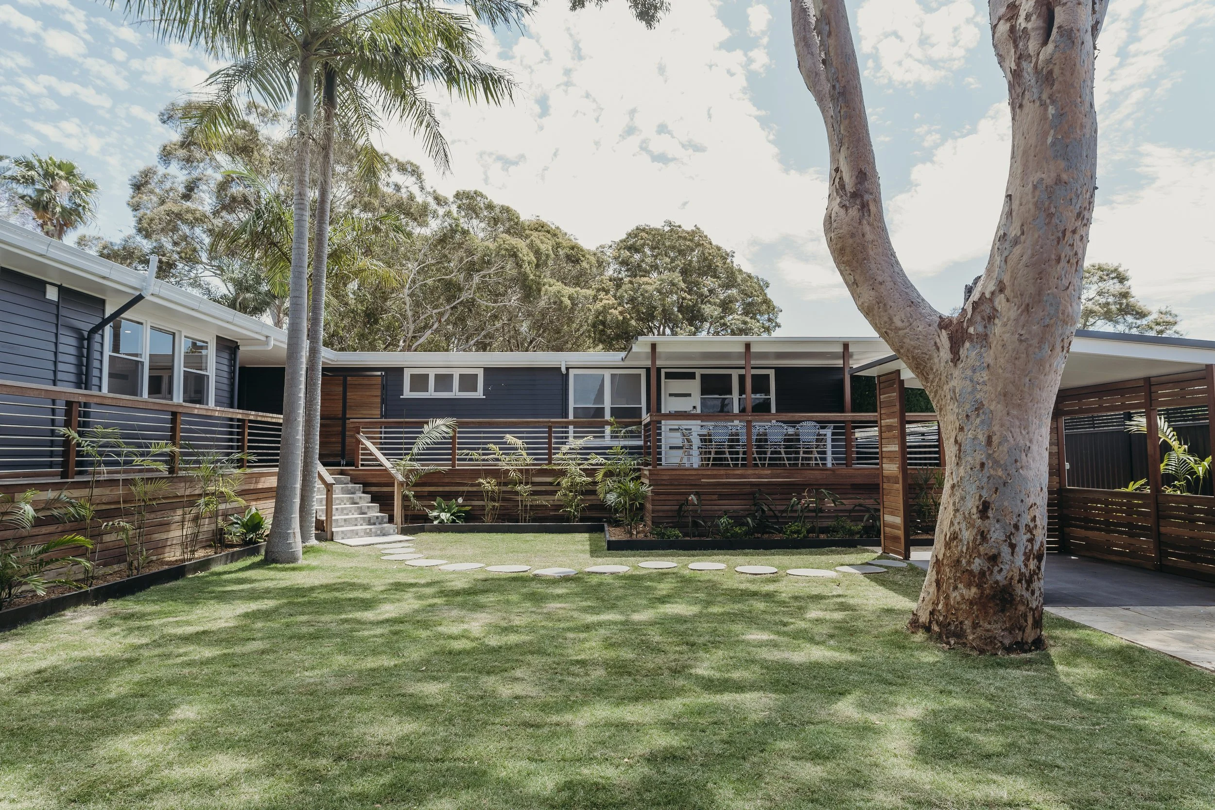 Modern house with a wooden deck, surrounded by a well-maintained lawn, trees, and tropical plants under a partly cloudy sky.