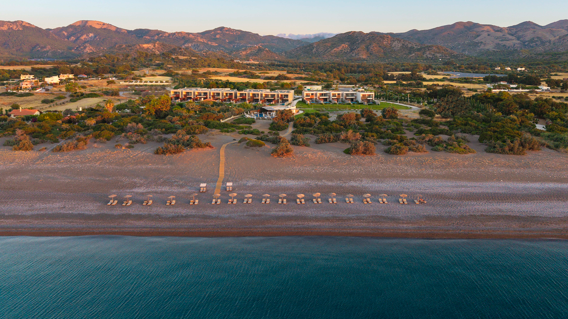 Rhodes Aerial view of a beachfront luxury resort in Rhodes, Greece, with sun loungers on a sandy beach, lush greenery, and mountains in the background
