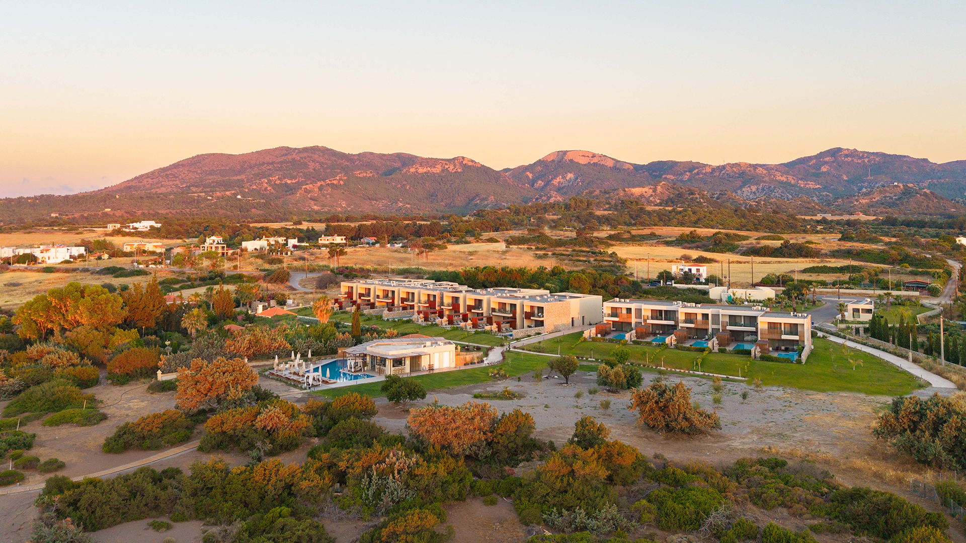 Rhodes Villas, lush greenery, swimming pools, and mountains in the background during sunset.