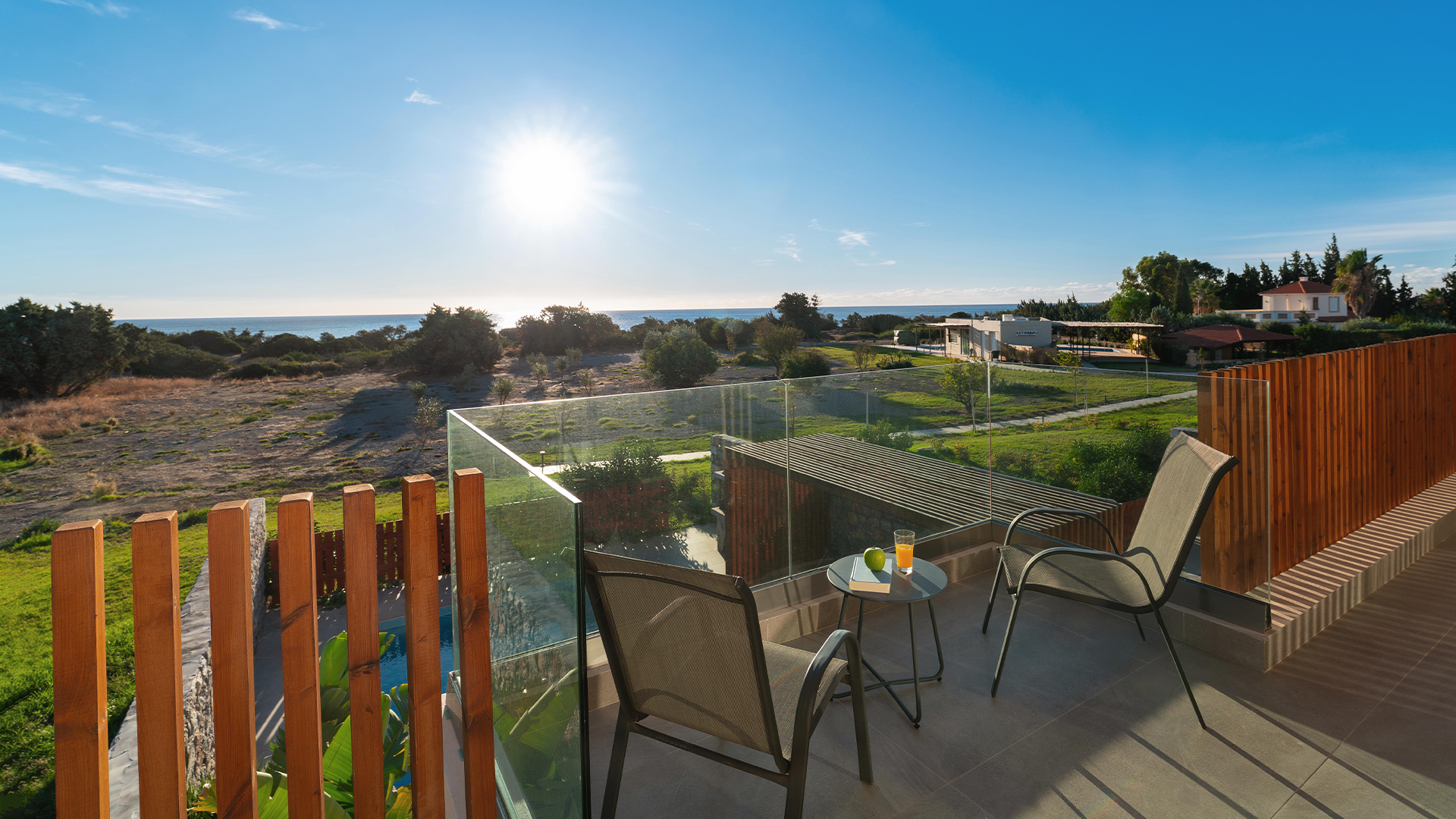 Rhodes Sea View Villa outdoor seating with overlooking a scenic landscape with trees, fields, and the ocean under a clear blue sky with the sun shining.