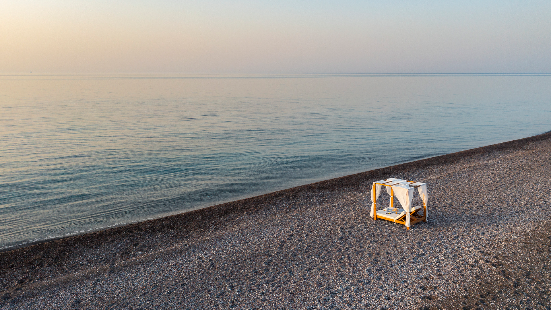 A small bed with white linens and a canopy, set up on a pebbled beach near calm water during sunset or sunrise.