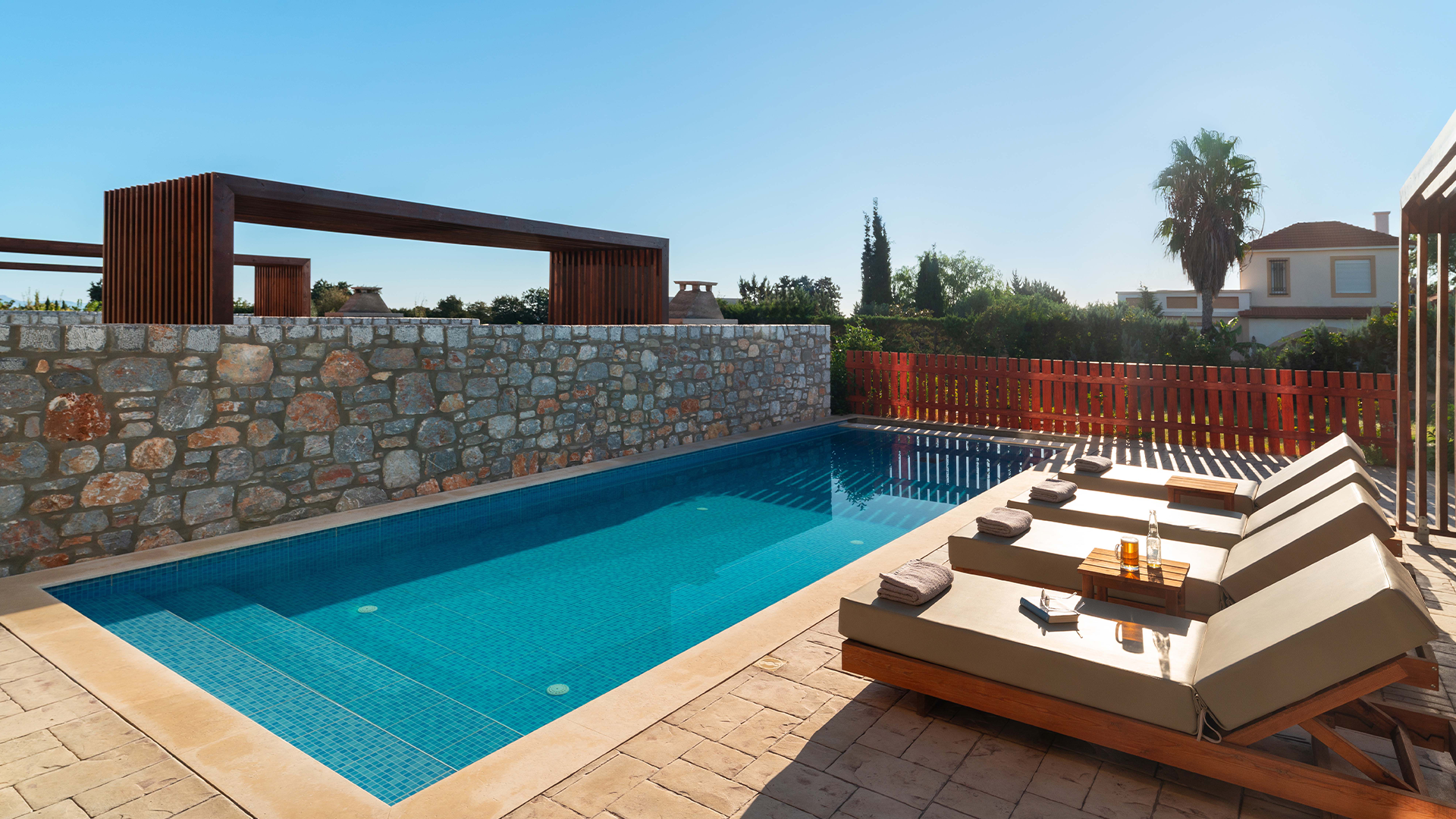 Rhodes Villa Outdoor swimming pool with lounge chairs, greenery and houses in the background under a clear blue sky.