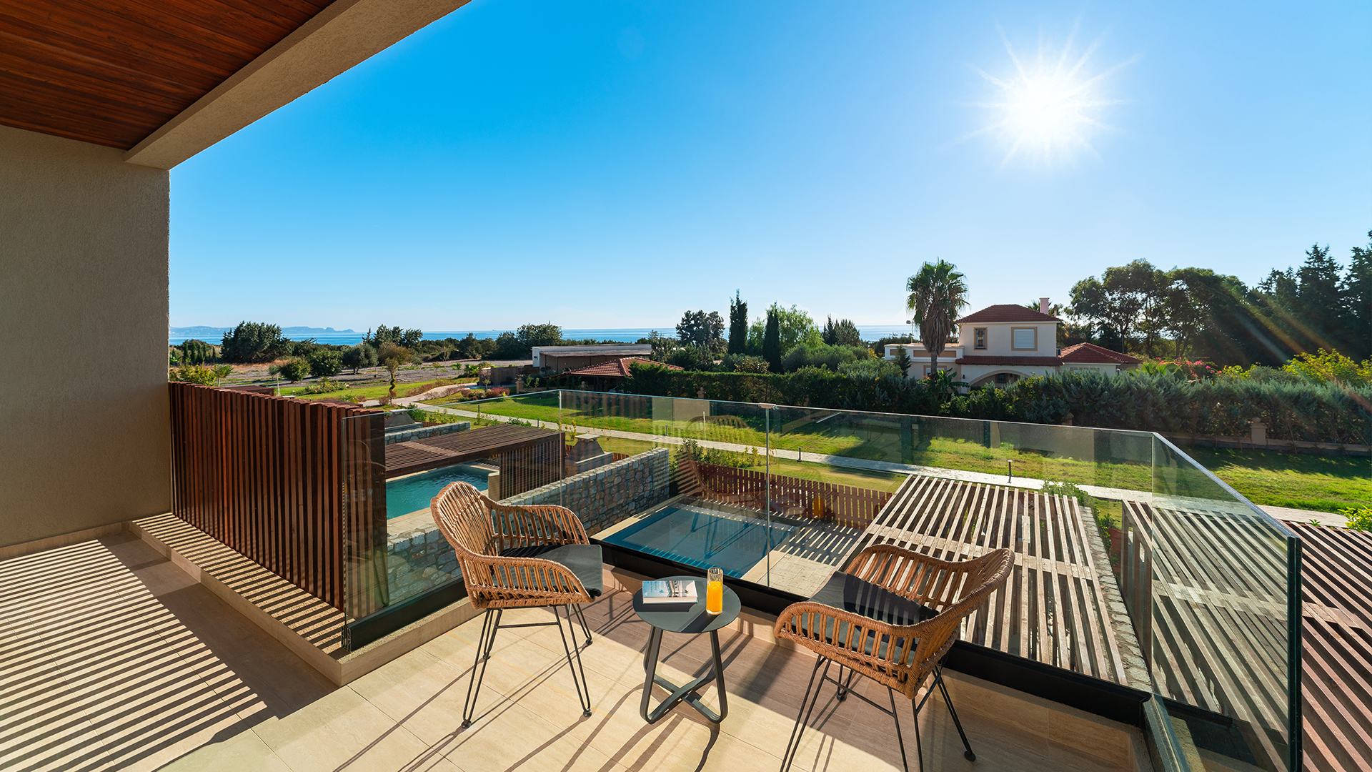 Rhodes Villa Balcony, overlooking a backyard with a pool, garden, trees, and a house under a bright blue sky with the sun.