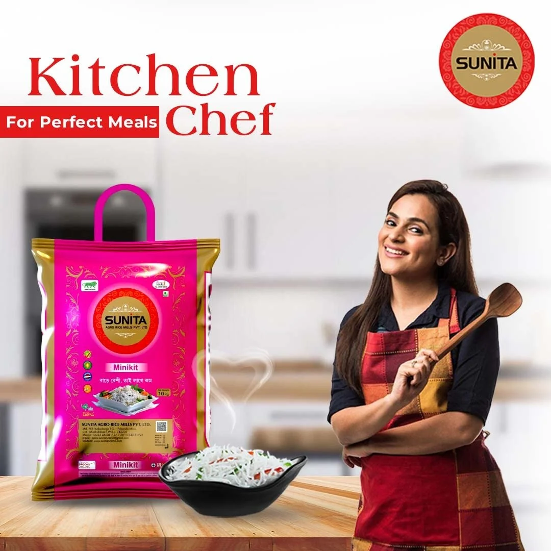 A smiling woman in a kitchen holding a wooden spoon with a package of Sunita Minikit rice and a bowl of rice noodles on a wooden table.