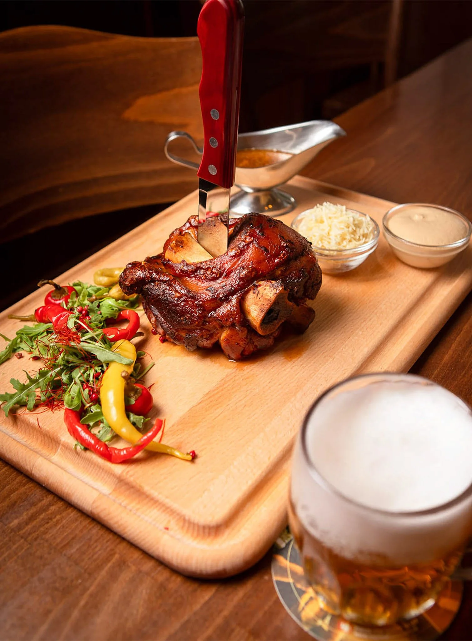 Roast pork knee with side salad of mixed greens and peppers, sliced cheese, sauces, and a Czech beer in a glass.