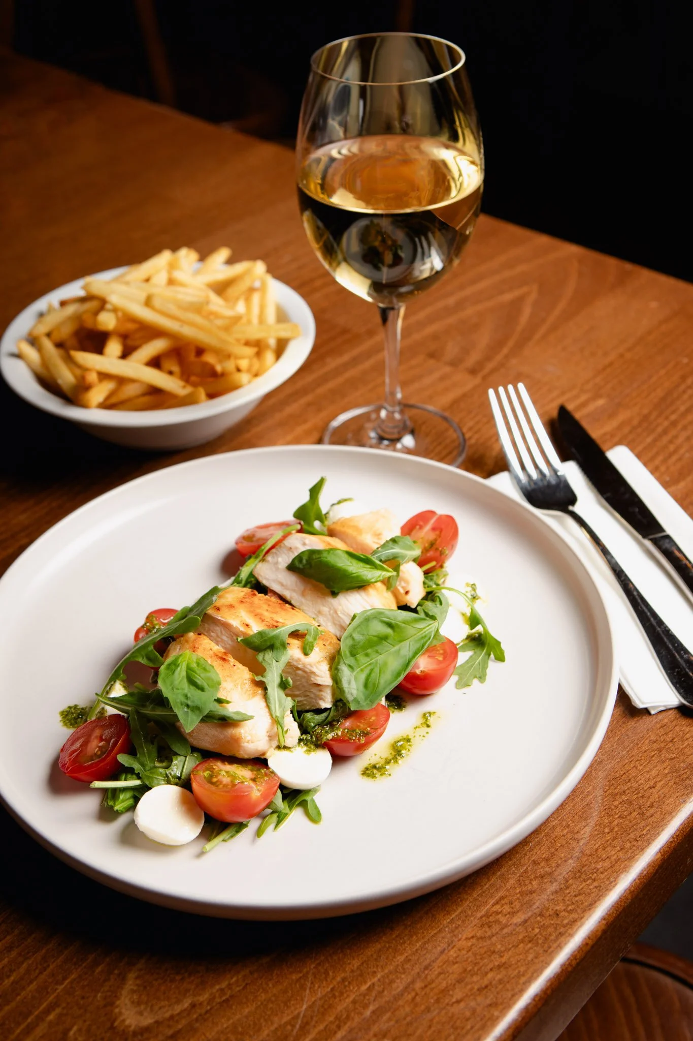 Grilled chicken steak with cherry tomatoes, mozzarella, and greens, alongside a side of French fries, a glass of white wine.