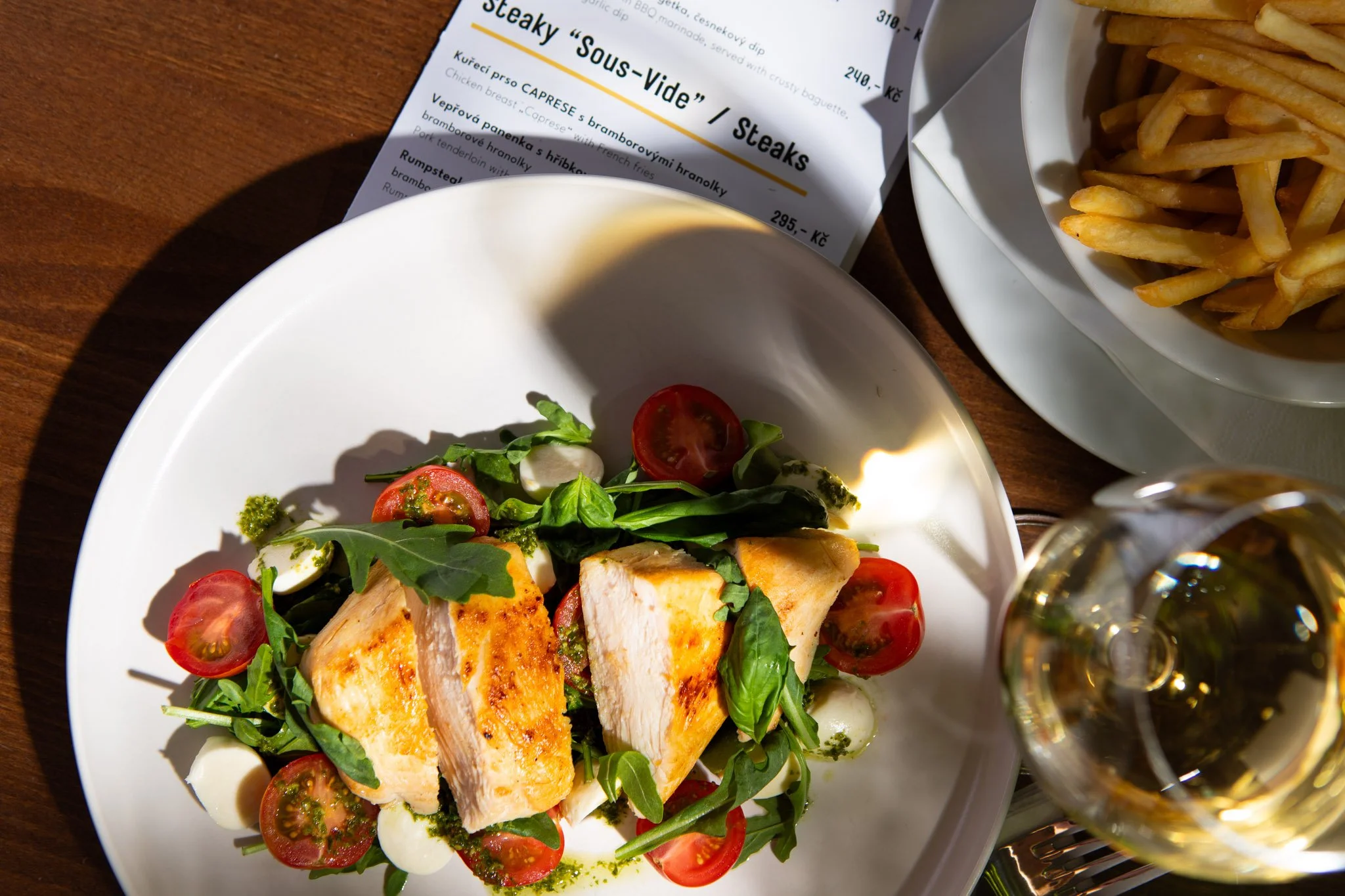 Grilled chicken steak with cherry tomatoes, mozzarella, and greens, alongside a side of French fries, a glass of white wine.