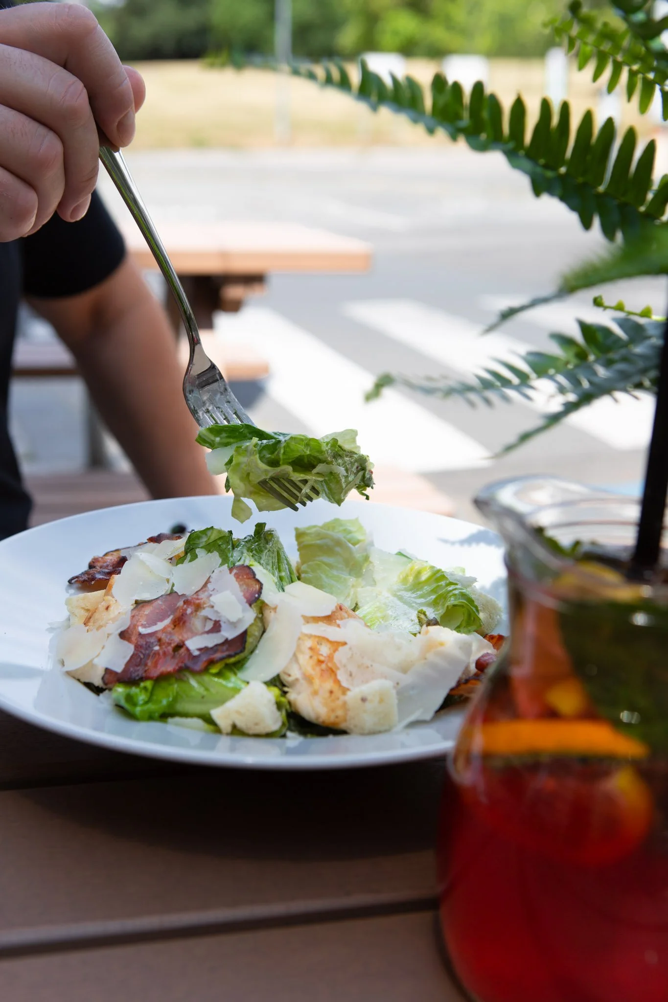 Person eating a salad with lettuce, bacon, cheese, and croutons at an outdoor restaurant table with a glass of iced tea in the foreground and greenery in the background.