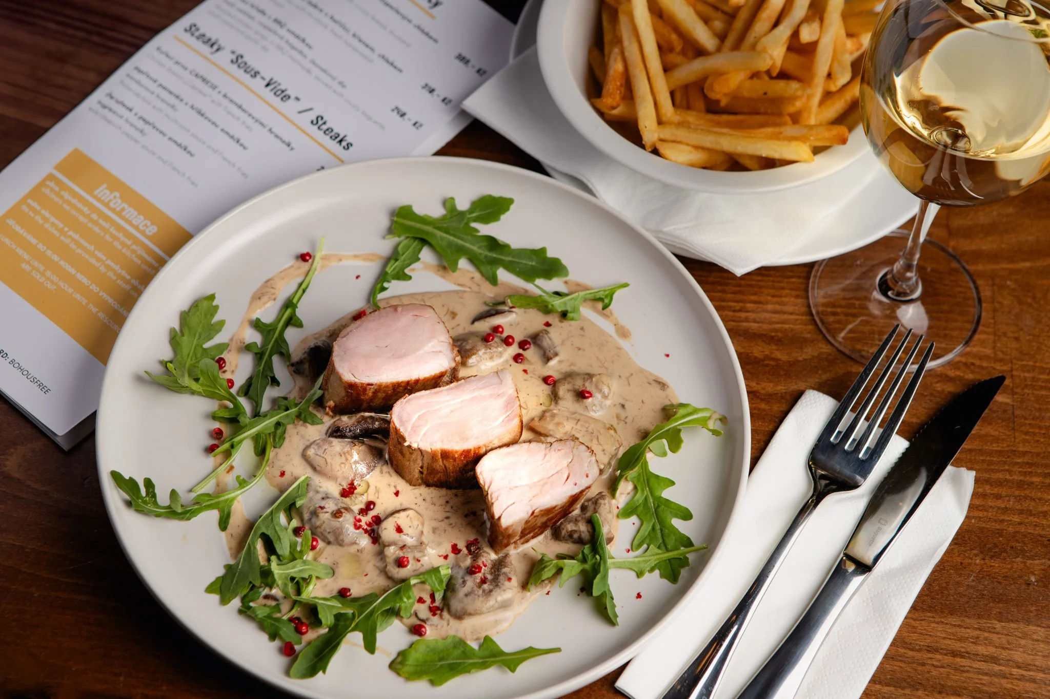 Sliced pork tenderloin topped with mushroom gravy, garnished with arugula and red peppercorns, accompanied by a bowl of French fries, a glass of white wine, Czech restaurant.