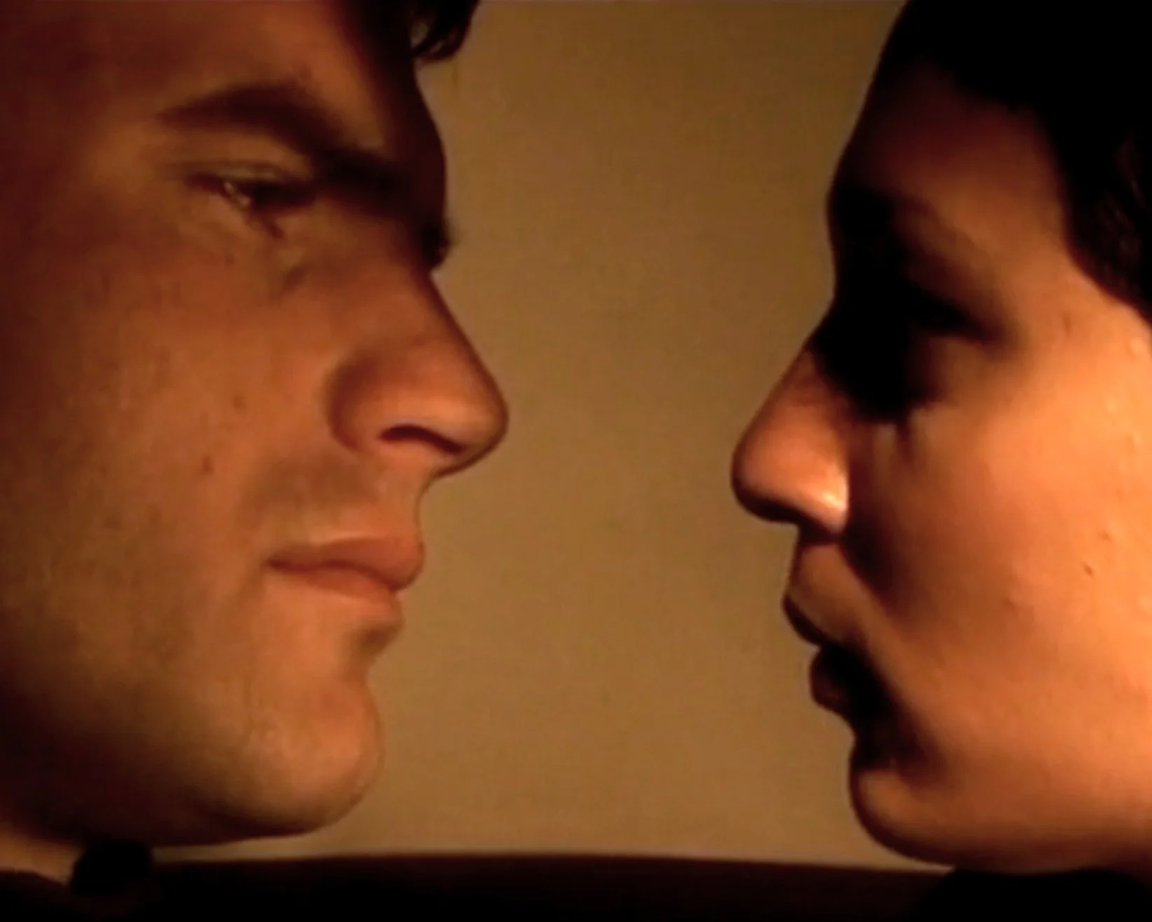 Close-up of two people, one with light skin and dark hair on the left, and one with slightly darker skin and dark hair on the right, facing each other with foreheads almost touching in an intimate moment.