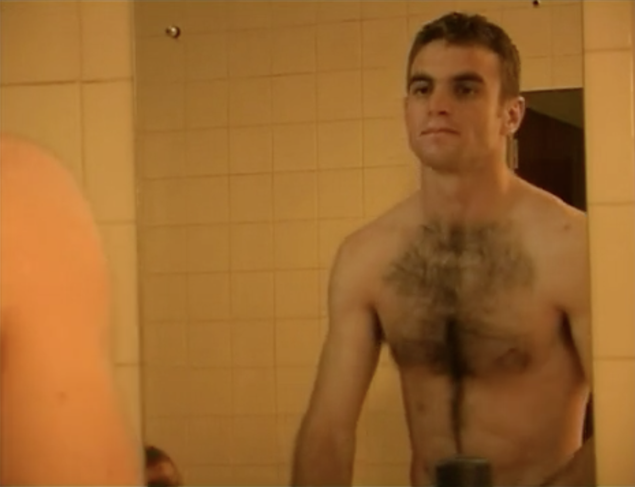 A shirtless young man with short dark hair and a beard stands in a shower area with yellow tiled walls.