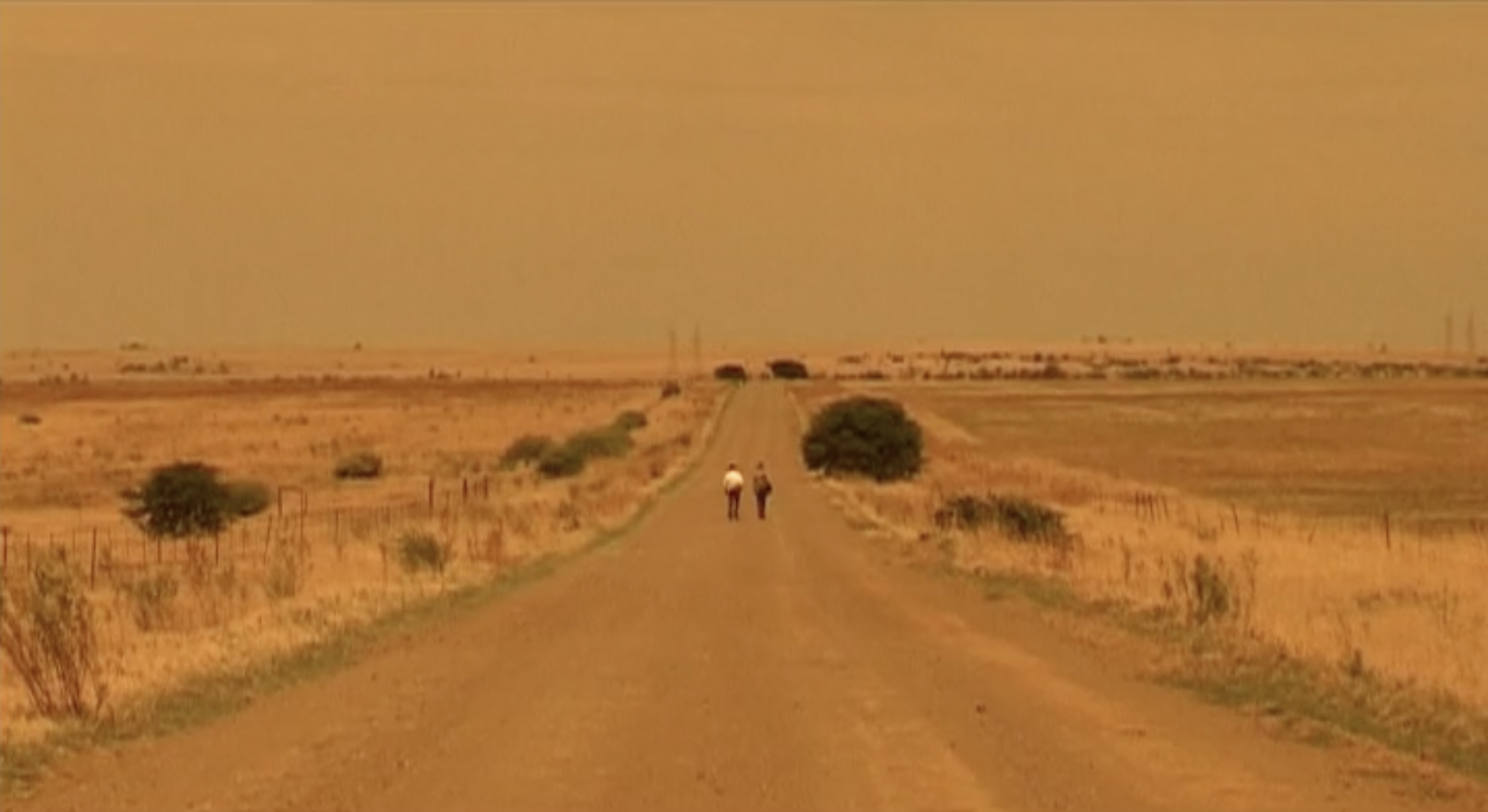 A straight dirt road running through a dry, flat landscape with sparse vegetation. Two people are walking along the road, and the sky is orange, suggesting a sunset or dust-filled atmosphere.
