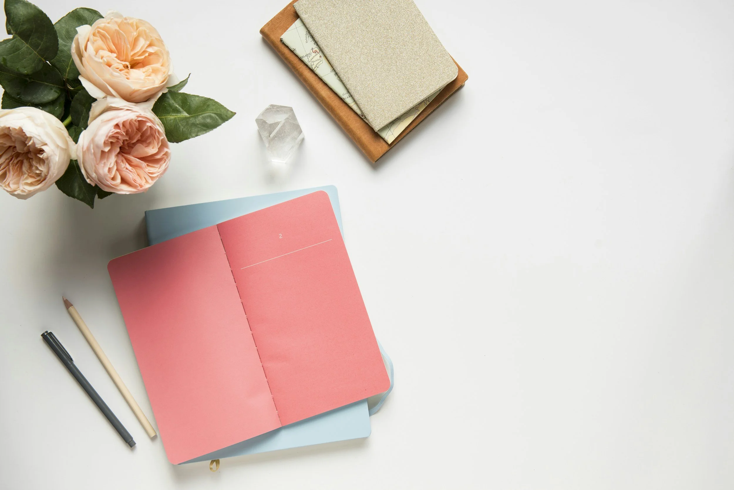 A flat lay of a white desk with a bouquet of pink roses, an open pink planner, a closed blue planner, black and beige pens, a small crystal, and a tan notebook with papers inside.