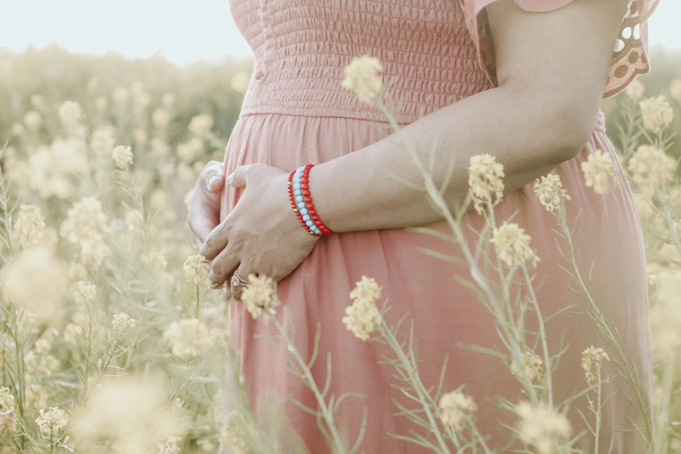 A person in a peach-colored dress stands in a field of yellow flowers, with their hands gently clasped around their abdomen, wearing red and blue beaded bracelets.
