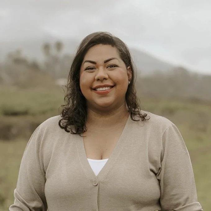 A woman smiling outdoors in a beige sweater with a white shirt underneath, standing in a natural landscape with mountains and cloudy sky in the background.