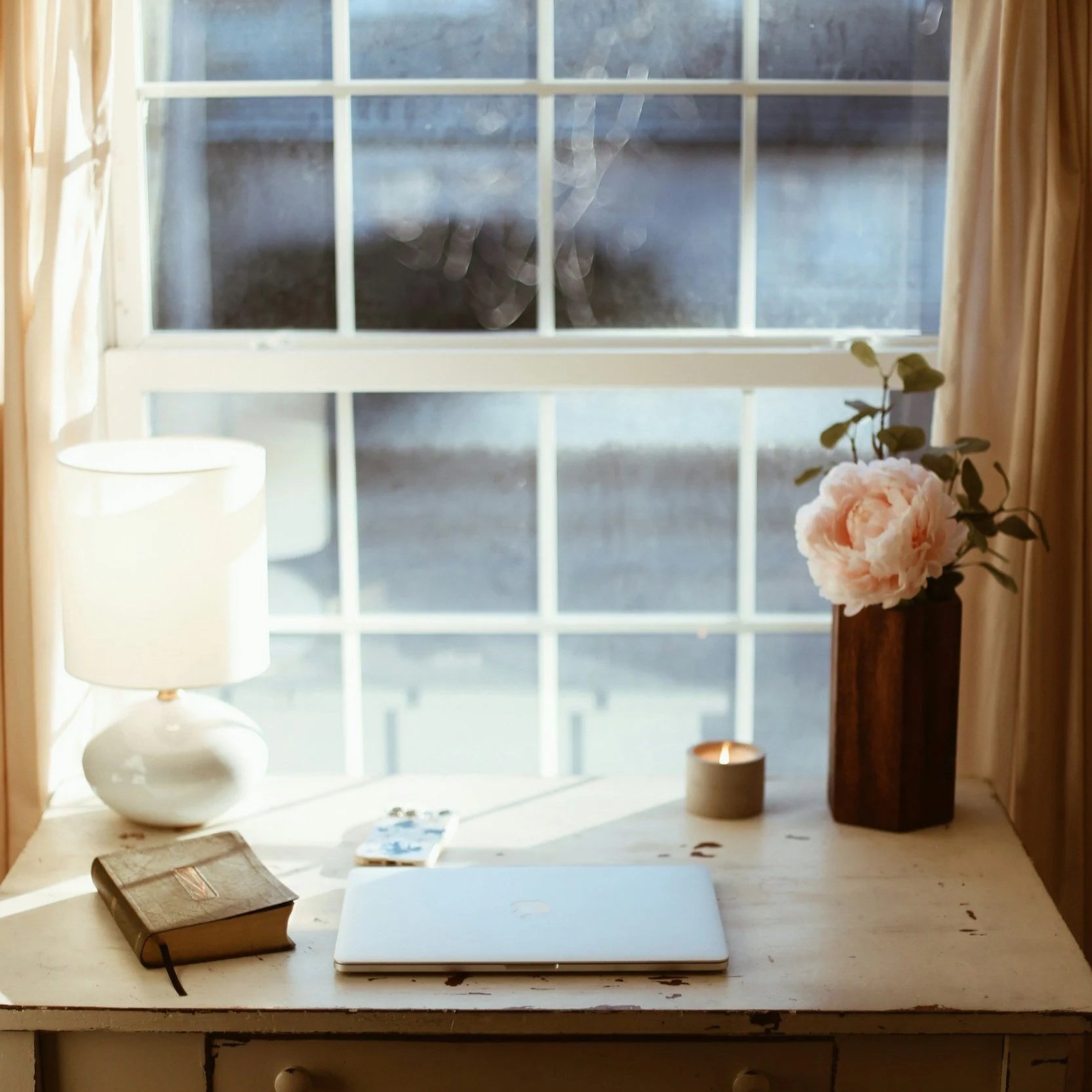 A white desk decorated with a closed silver laptop, a small scented candle, a brown journal, and a stack of small photographs or postcards. To the right, there is a tall dark wooden vase with white and pink peony flowers and green leaves. To the left, there is a white table lamp with a rounded base. The desk is near a window with white frame and divided panes, allowing sunlight to brighten the space. beige curtains frame the window.