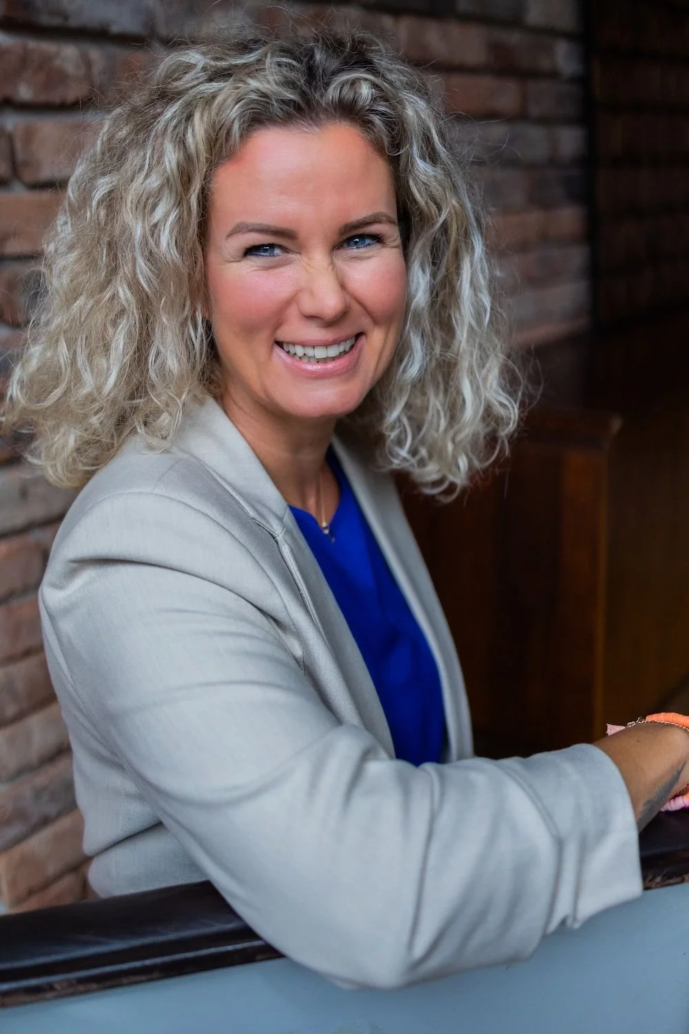Lächelnde Frau mit lockigem blondem Haar, trägt grauen Blazer und blaßes Oberteil, sitzt vor Backsteinmauer.