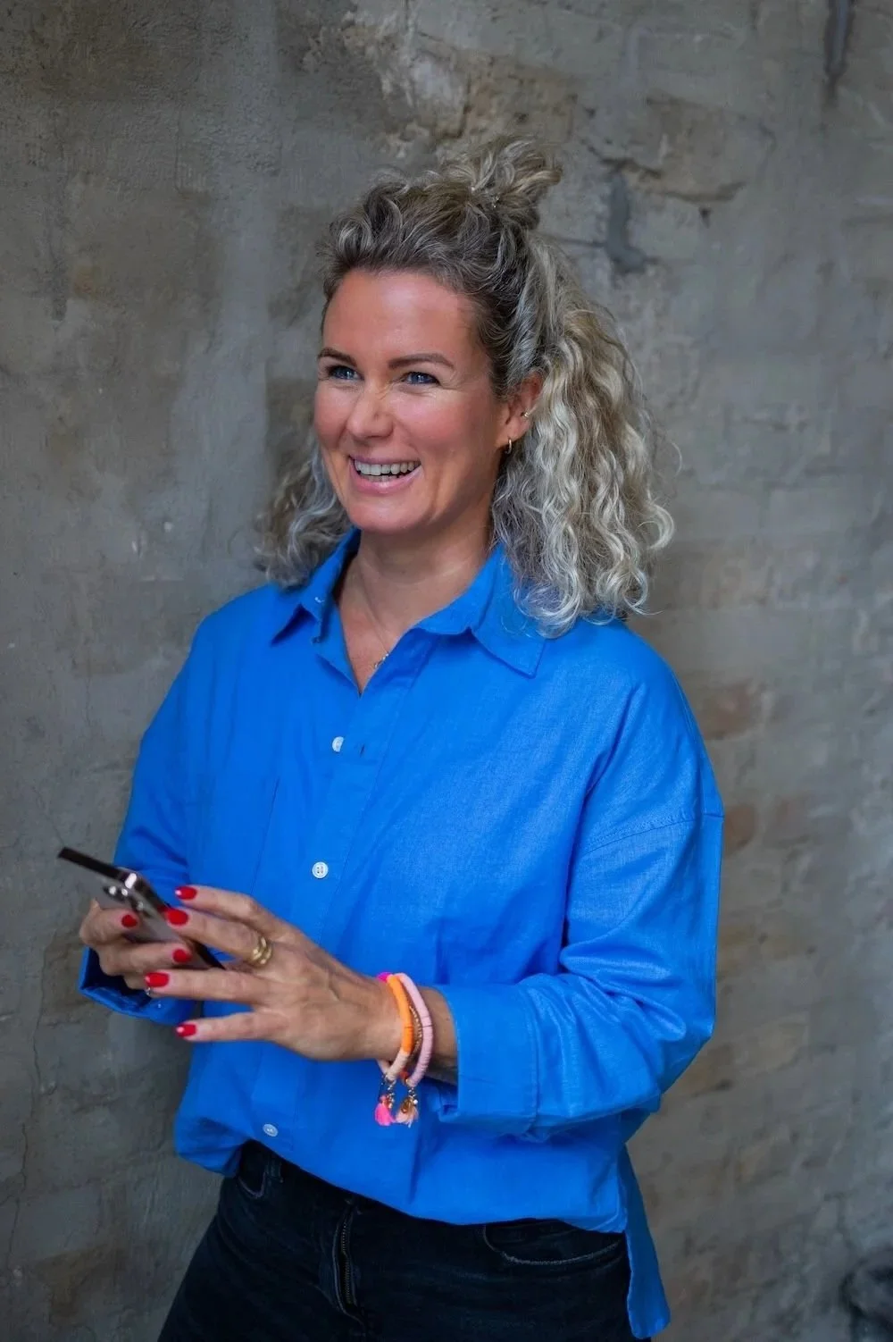 Felicitas Geppert, Ordnungscoach im Ortenaukreis, lachend in blauer Bluse mit Smartphone vor einer Steinwand