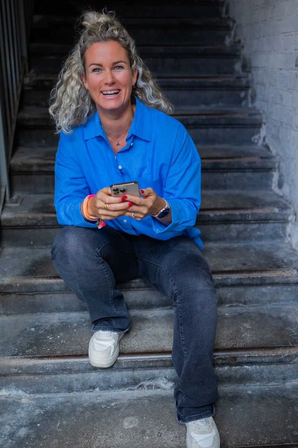 Eine lachende Frau mit lockigem grauem Haar sitzt auf einer steinernen Treppe und hält ein Smartphone in den Händen.