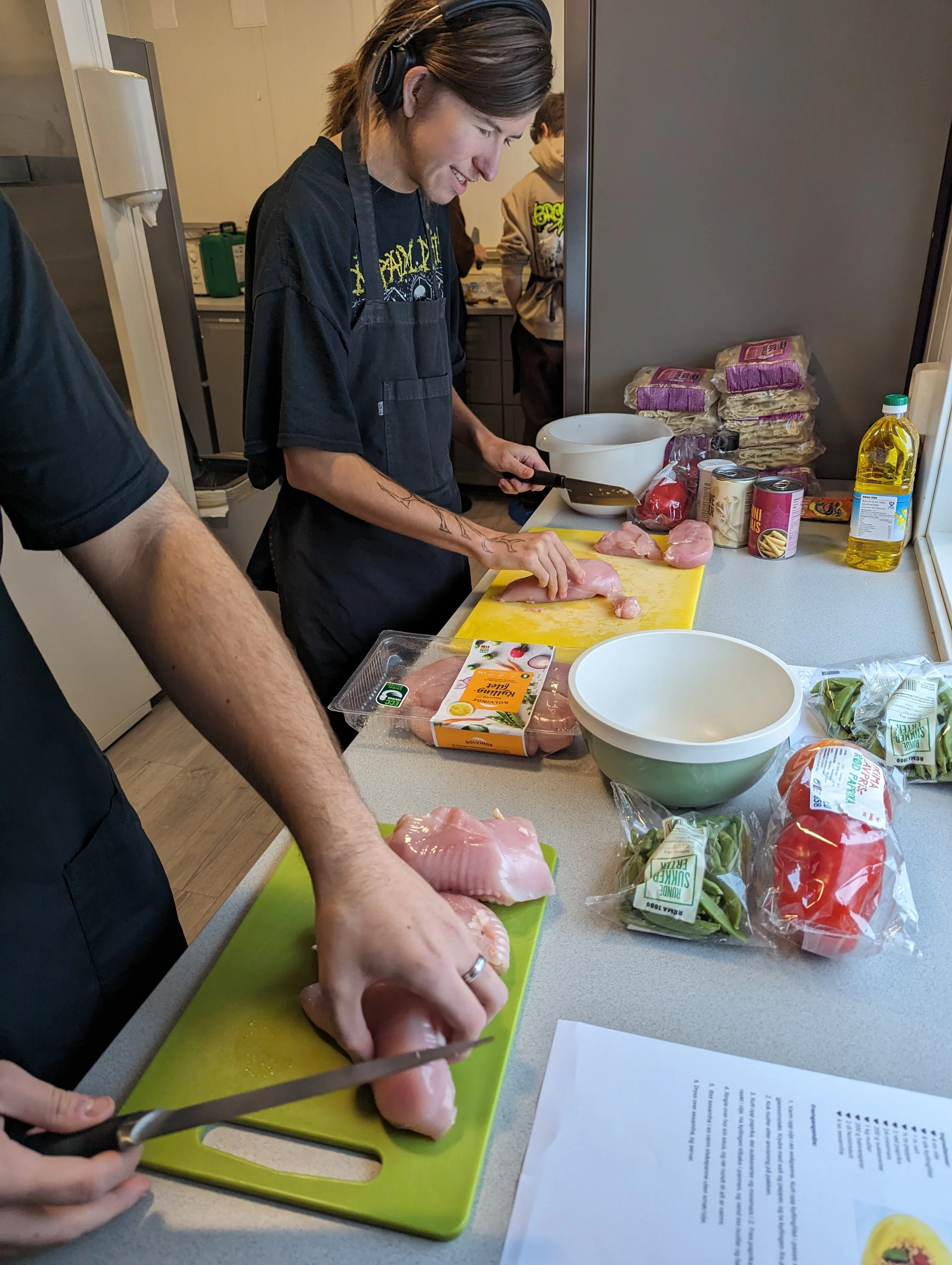 To personer som lager mat på et kjøkken, en kvinne som skjærer kylling på en grønn skjærefjøl og en mann som holder et stykke kylling i hånden. Kjøkkenbenken har pakker med kylling, grønnsaker, hermetikk og andre matvarer.