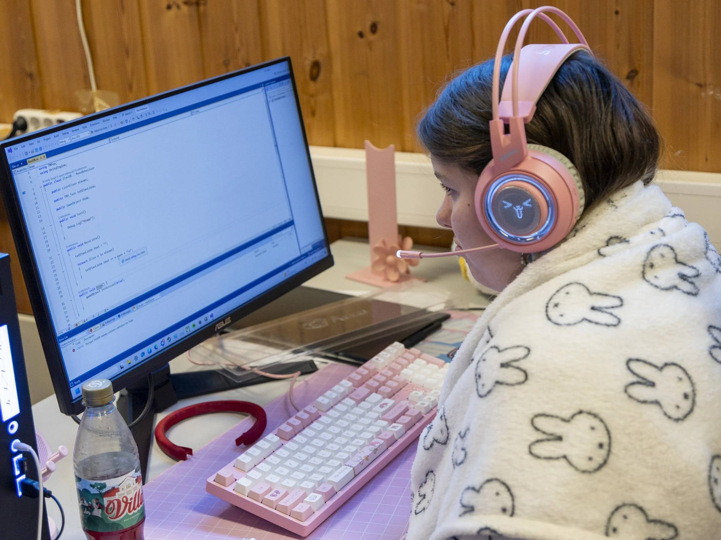En jente med rosa hodetelefoner og et hvitt skjerf med kaninmotiver, som jobber med programmering på en datamaskin.