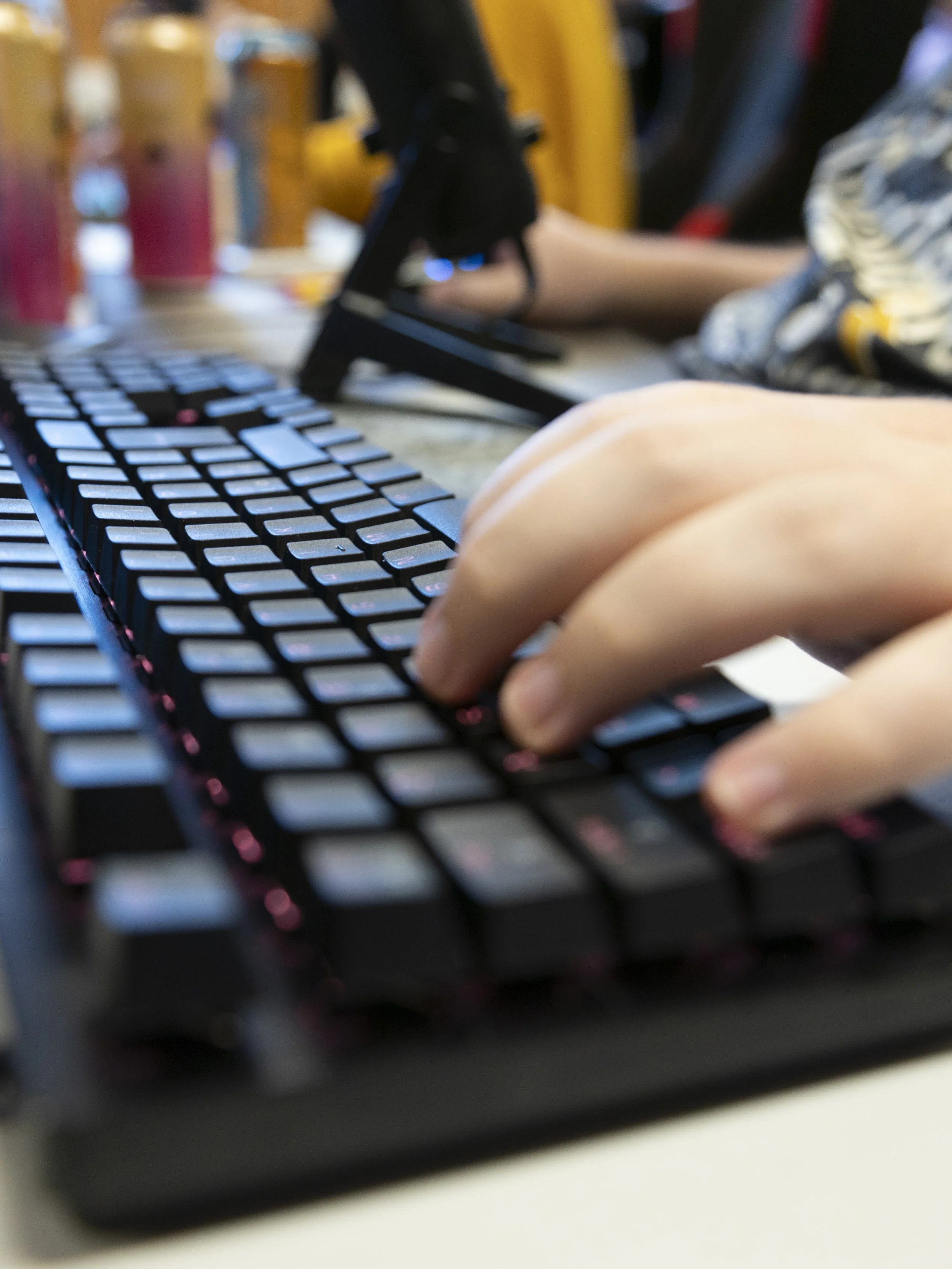 Nærbilde av en hånd som skriver på et svart tastatur med rosa bakgrunnsbelysning, med noen utstyr og gjenstander i bakgrunnen.